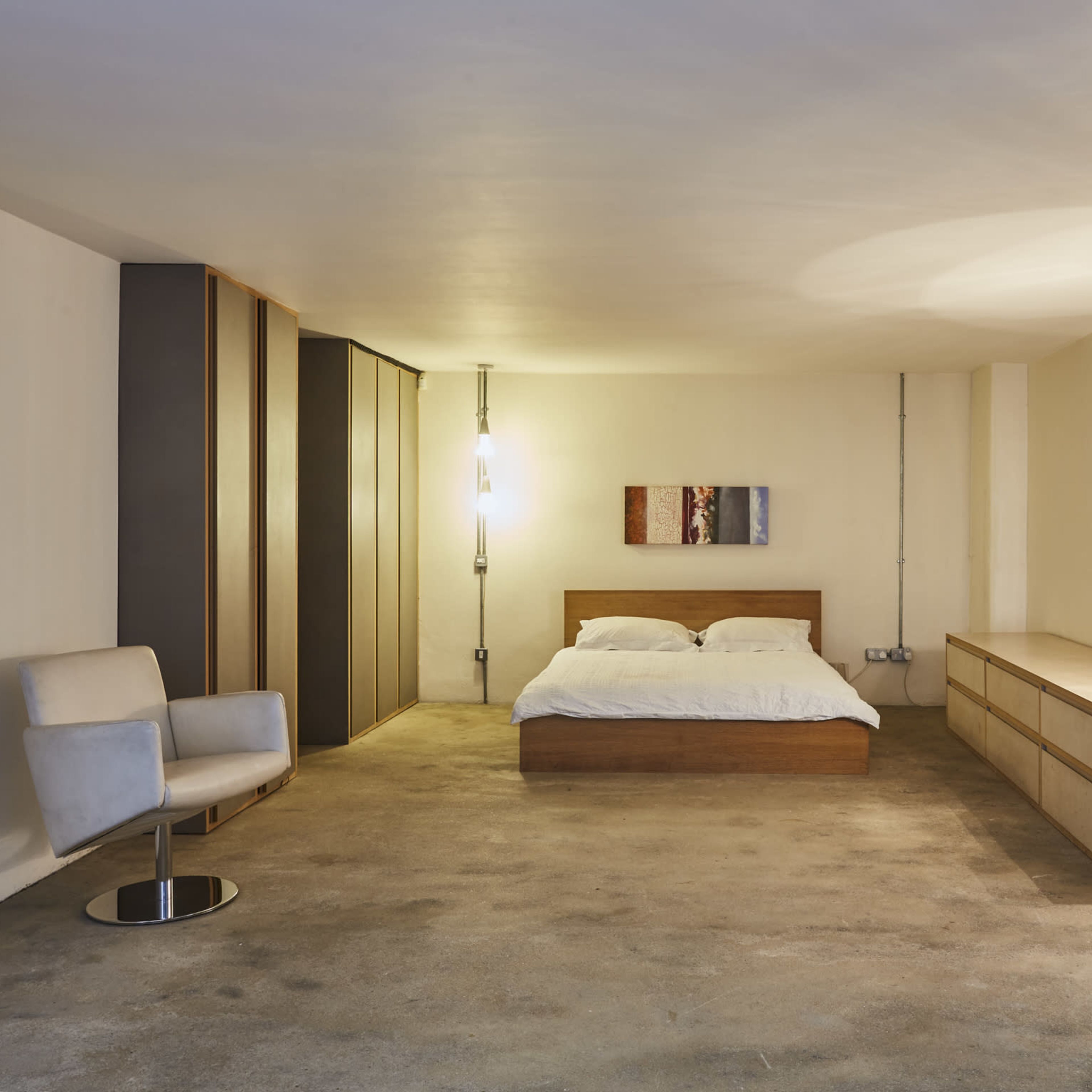 A minimalist bedroom with a wooden bed, a light-colored dresser, a modern chair, and soft lighting.