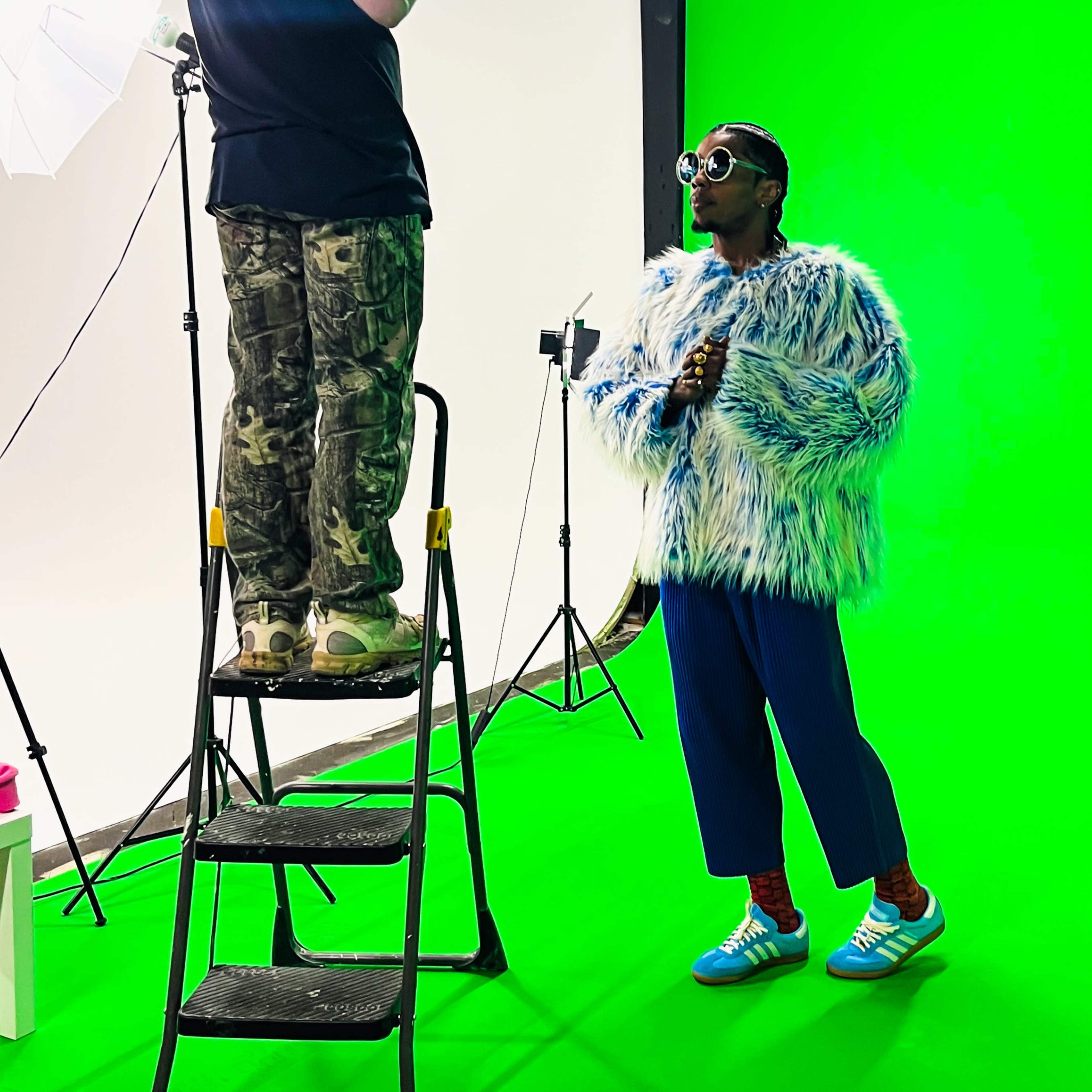 A split scene with four quadrants: a man speaking on camera in front of a green screen, a motorcycle stunt performer doing a trick, a large empty studio with white and green backdrops, and a man using a mobile phone while standing in front of a green backdrop.