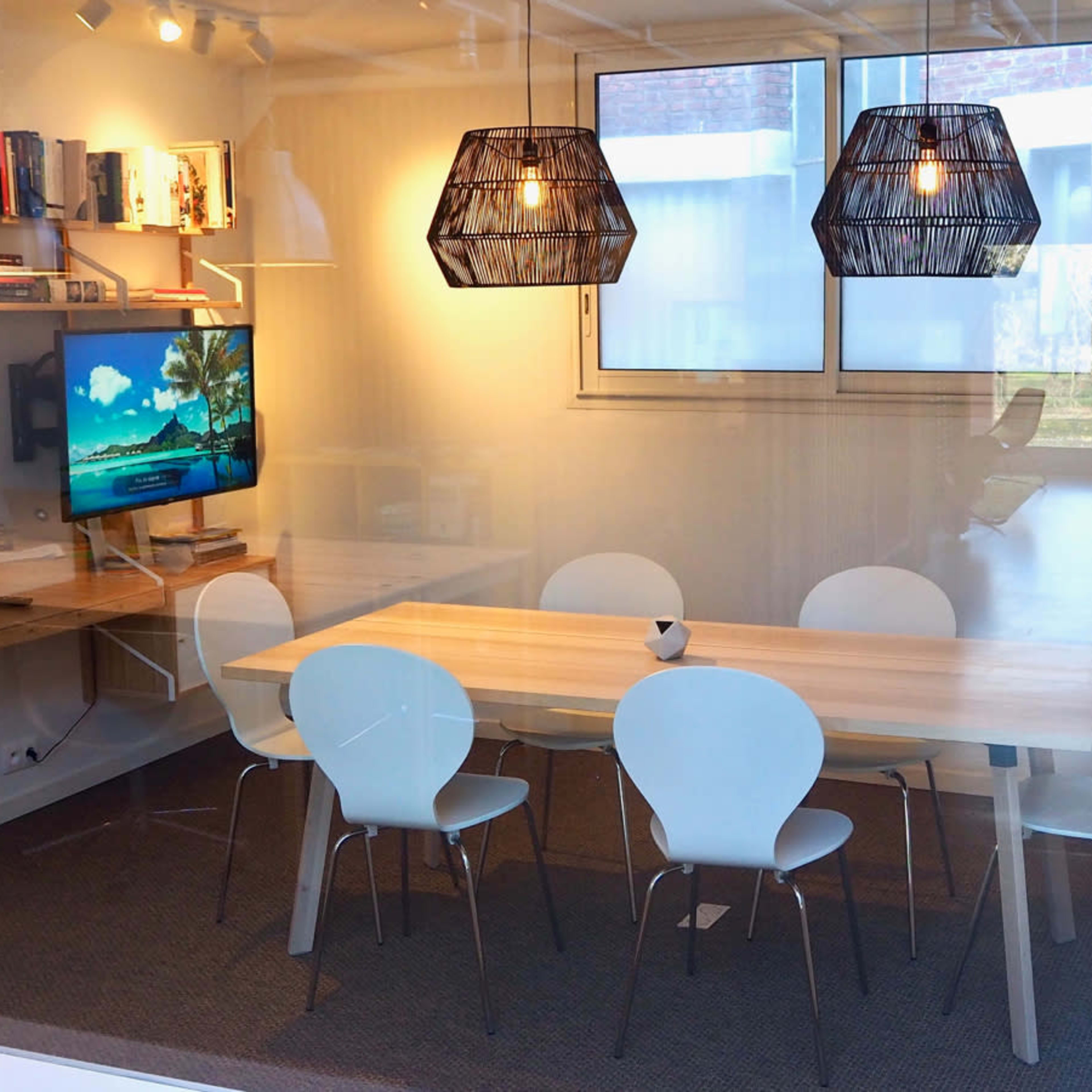 A modern conference room featuring a wooden table surrounded by white chairs, a wall-mounted television, and two pendant lights above the table.