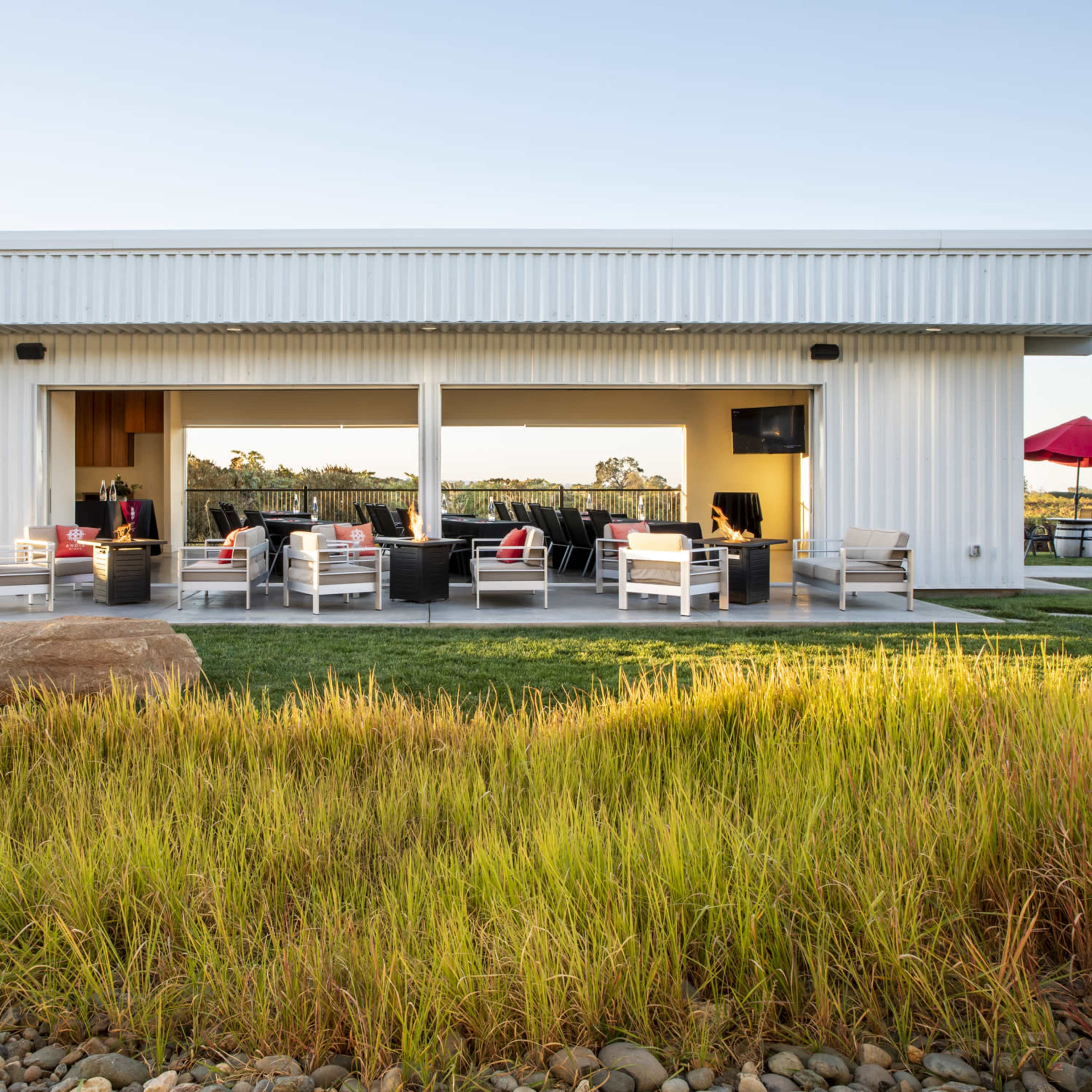 A modern, single-story structure with large openings and outdoor seating overlooks a grassy area and is set against a clear sky.