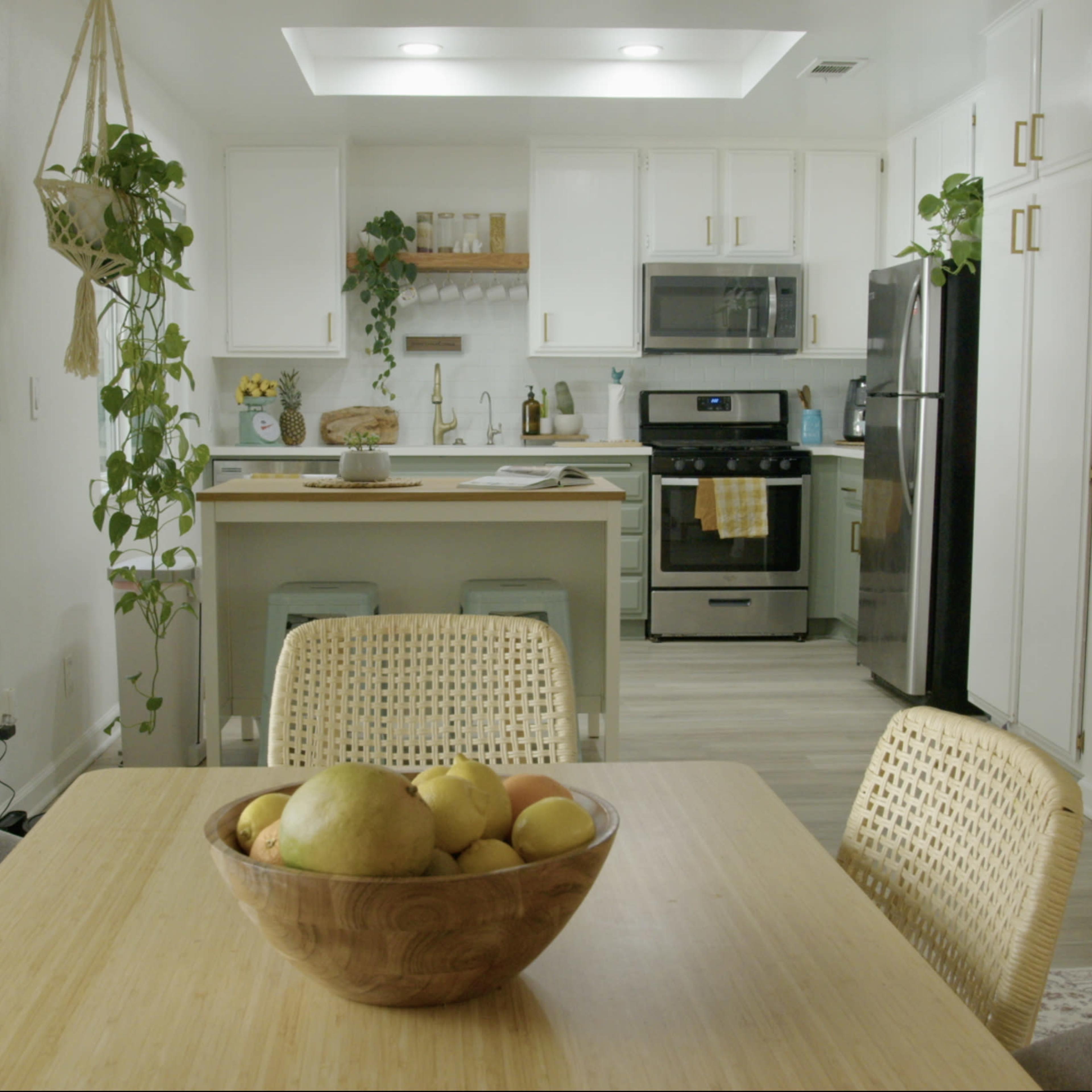 A kitchen and dining area feature a wooden table with a fruit bowl, surrounded by wicker chairs, while the kitchen has white cabinets and stainless steel appliances.