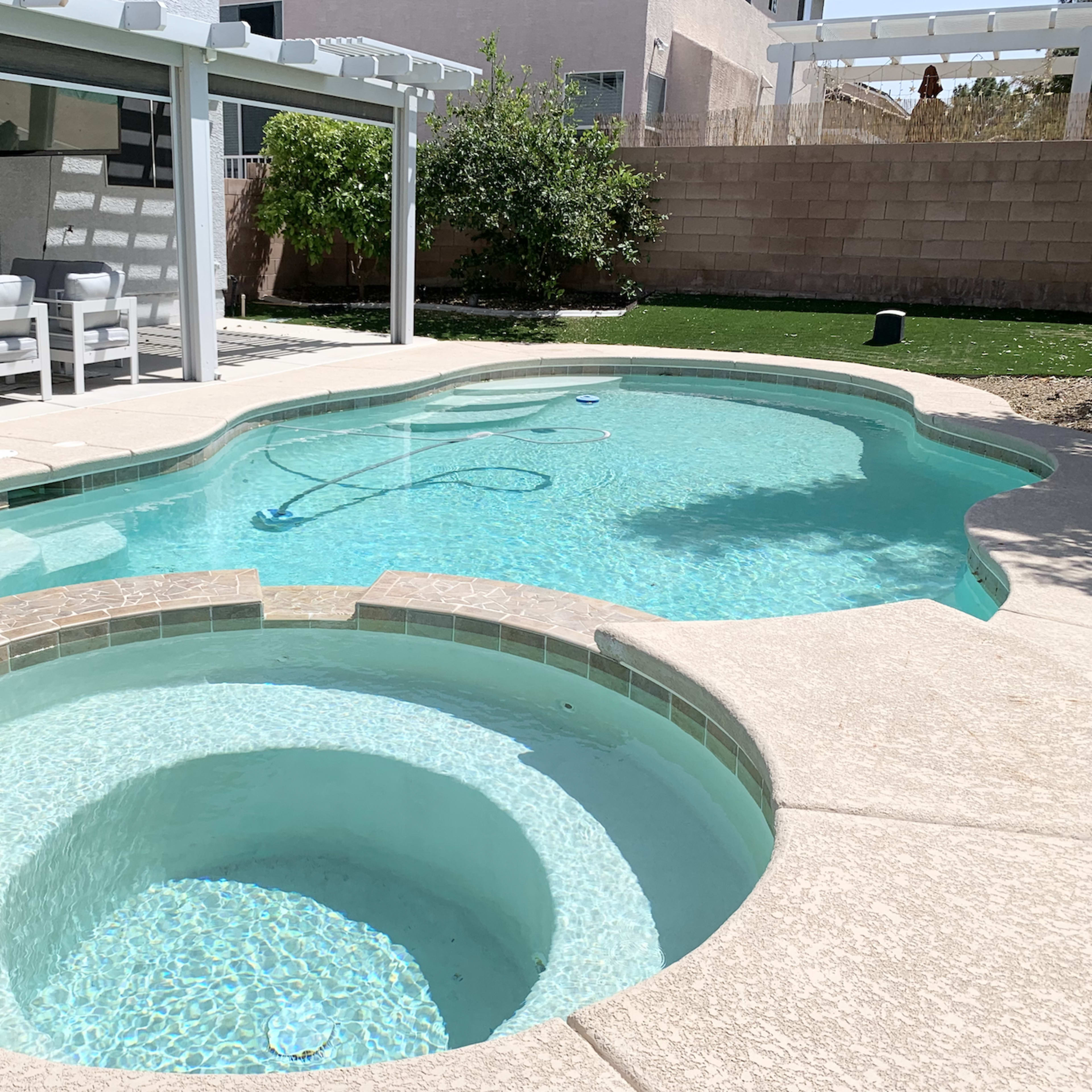 A backyard swimming pool with a connected spa, surrounded by a concrete patio and a shaded seating area.