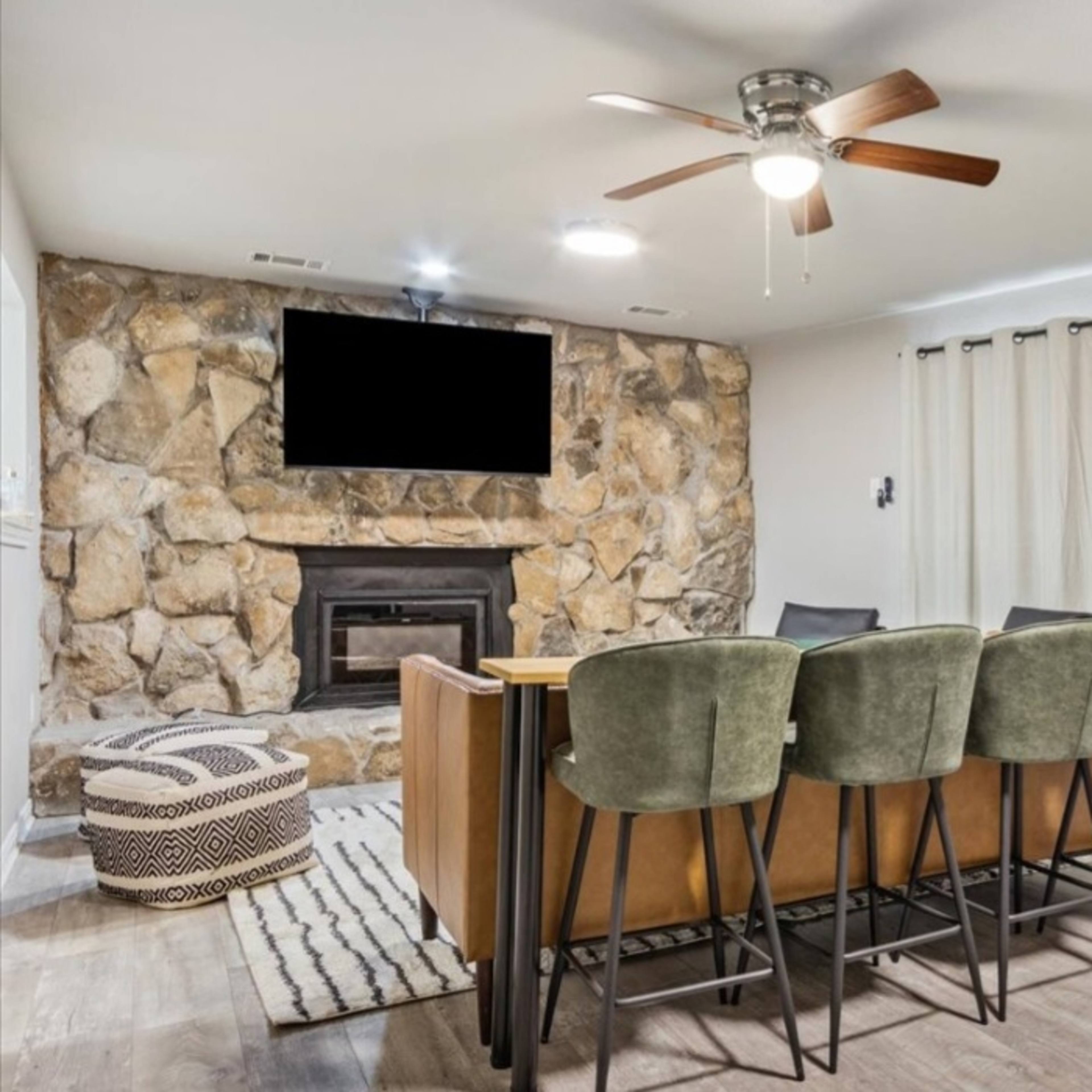 A living room with a stone fireplace, a television mounted above it, a dining table with high chairs, and beige curtains covering the windows.