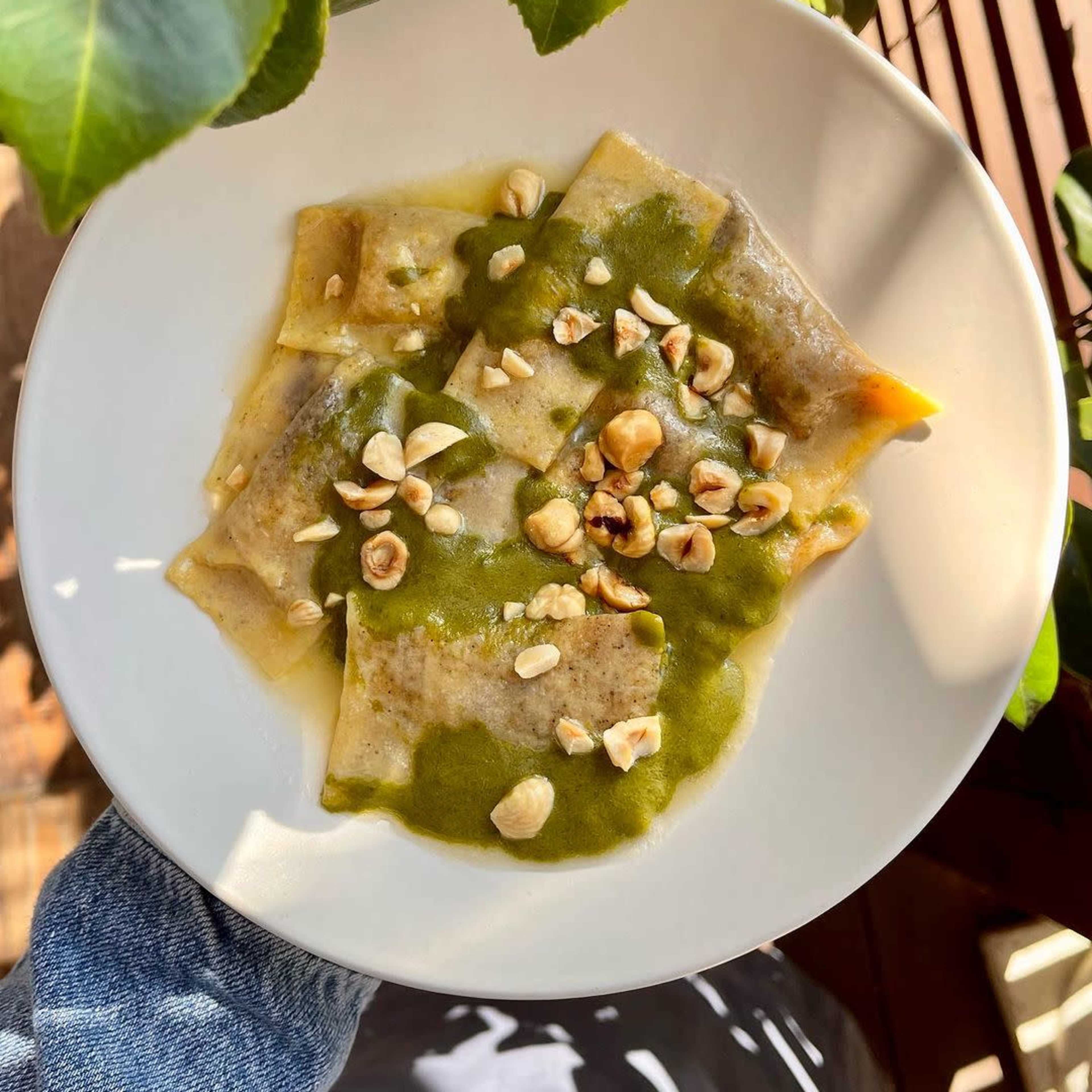 A plate of ravioli is topped with a green sauce and sprinkled with chopped nuts, set against a backdrop of leaves.