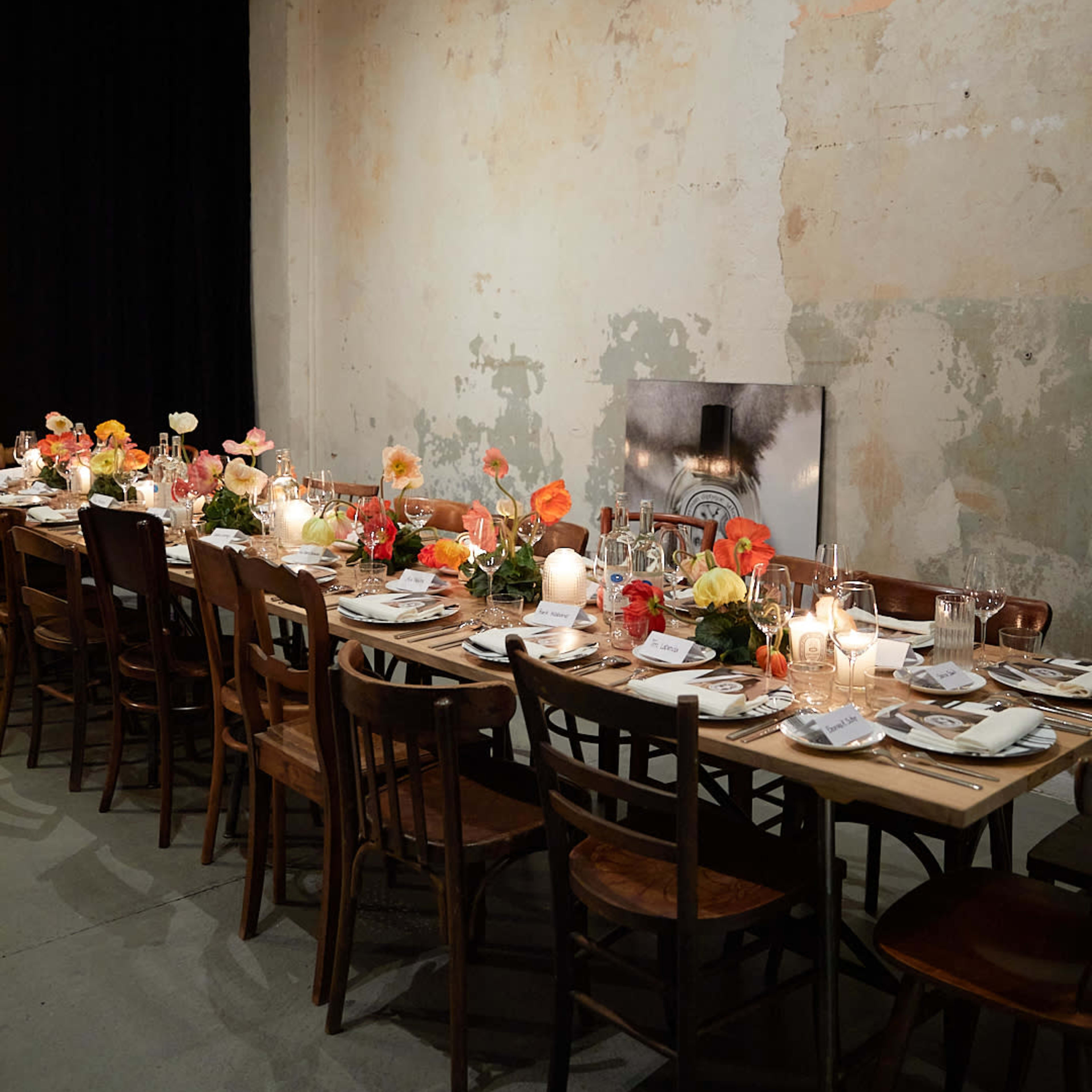 A long, elegantly set dinner table is adorned with colorful flowers and candles against a backdrop of a weathered, peeling wall.