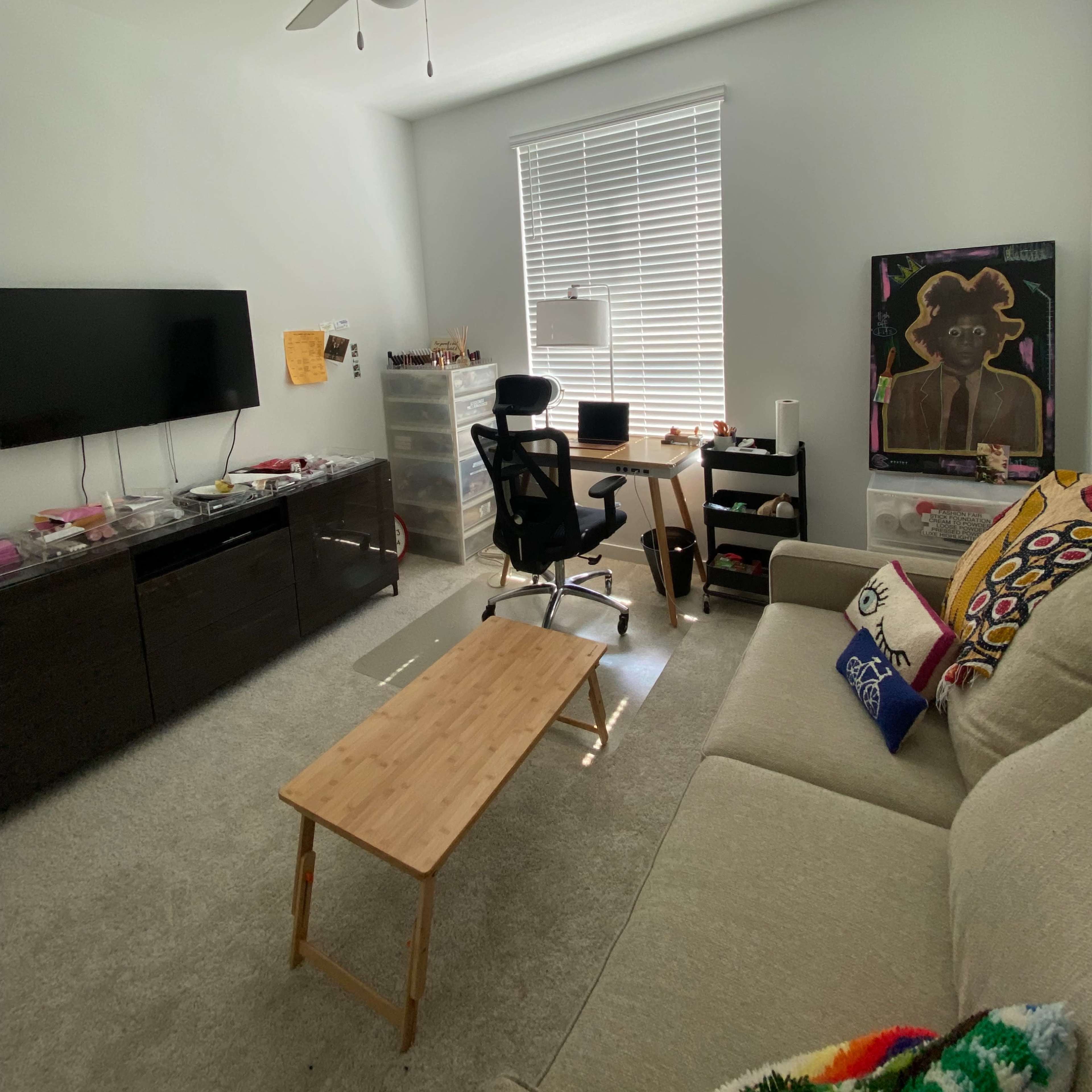 A living room with a sofa, a wooden coffee table, a desk with a chair, a television, and a large window with blinds.