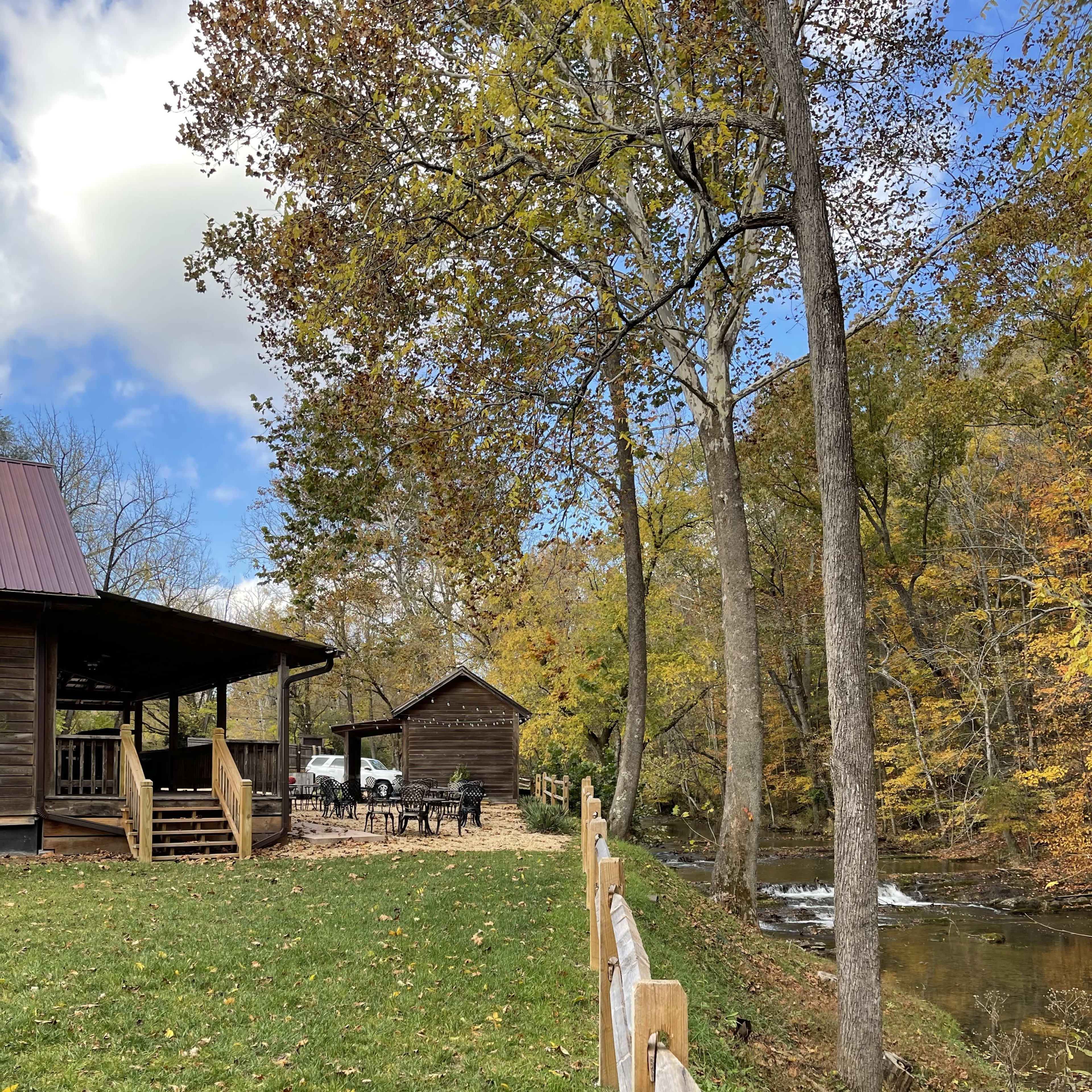 A rustic wooden cabin sits beside a gently flowing stream, with a grassy area and a wooden fence in the foreground.