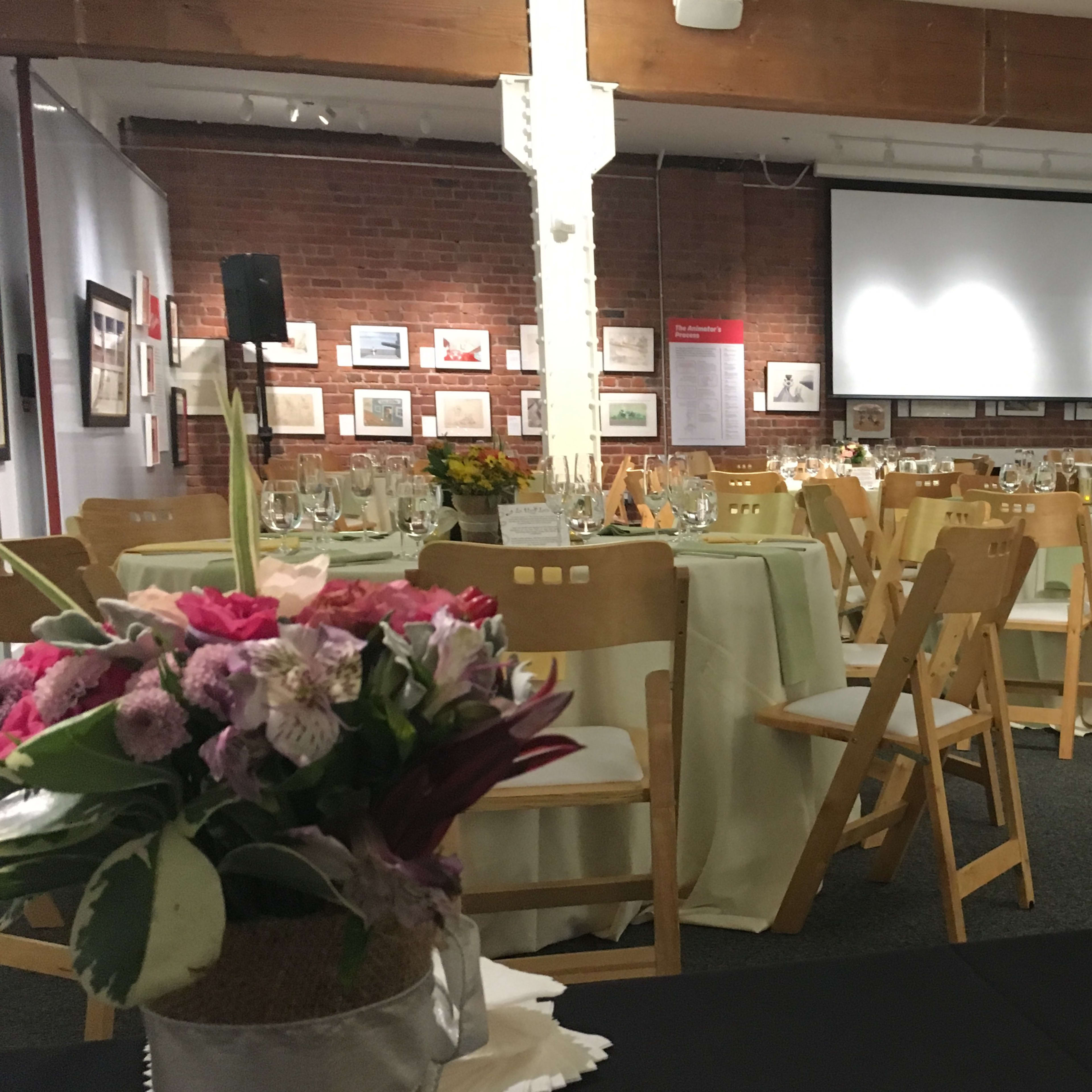 A dining setup is arranged in a hall with tables dressed in light-colored linens, featuring floral centerpieces and a projector screen in the background.