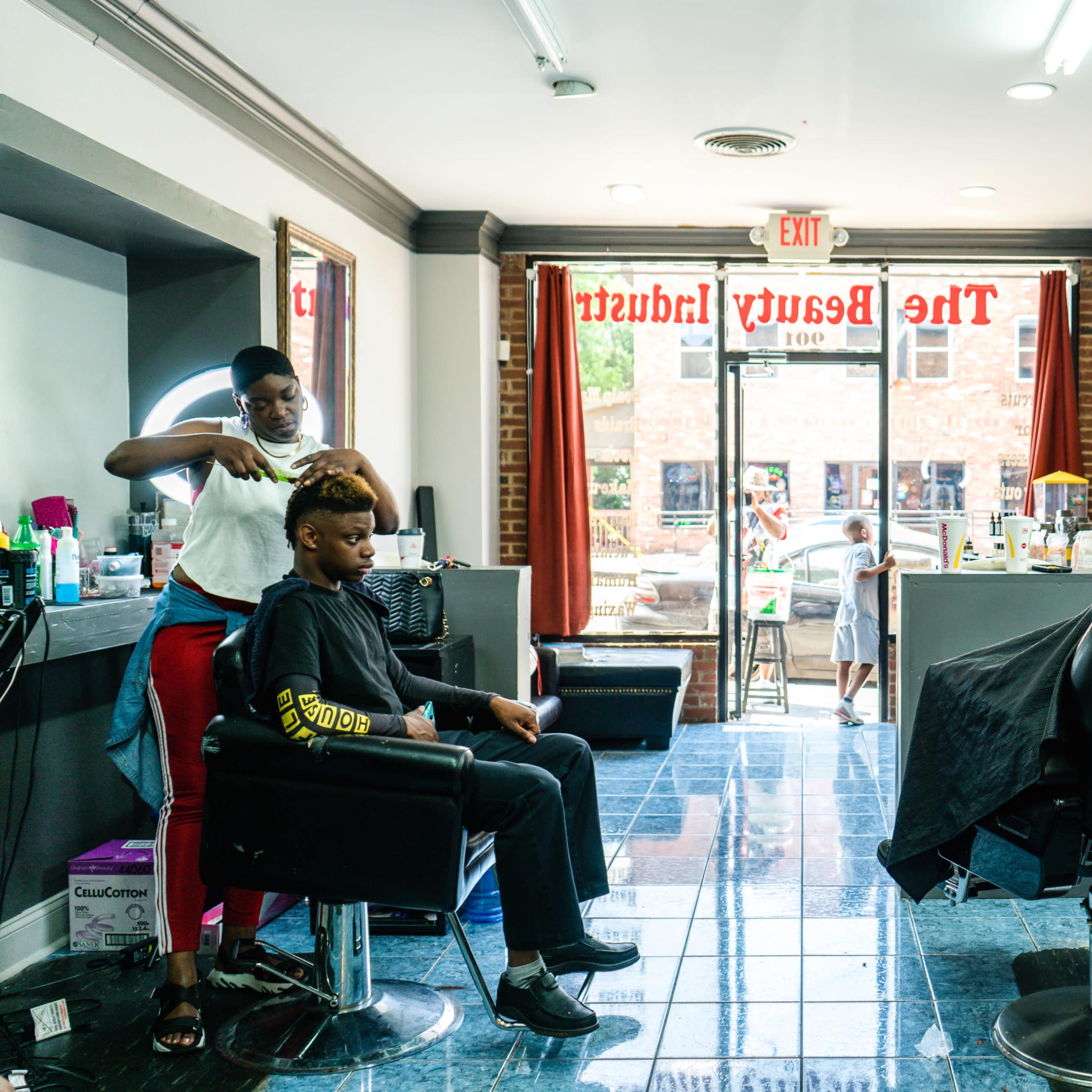 A barber shop interior features two clients in chairs, with one barber cutting hair and another stylist attending to a different client.