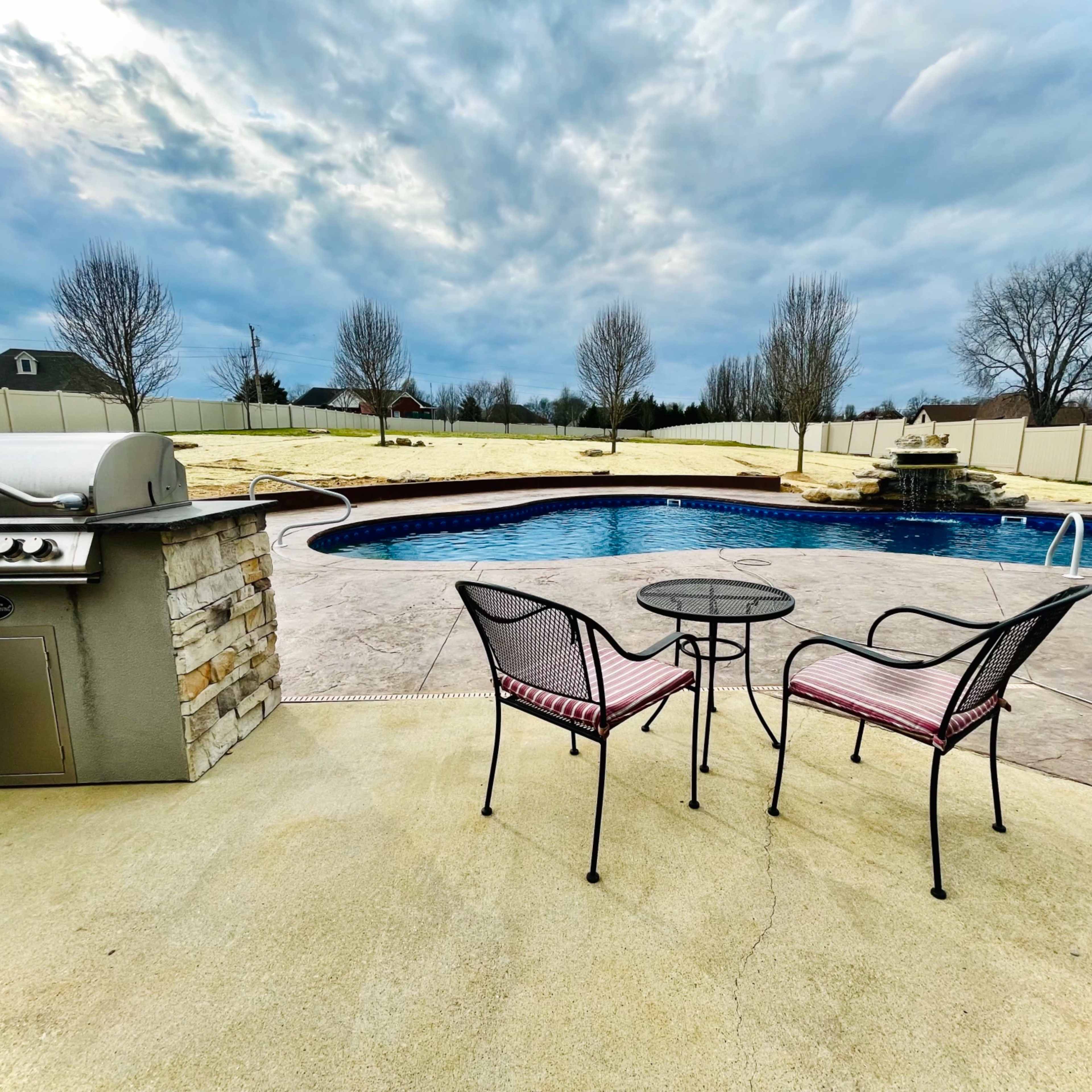 A poolside area with a grill, two chairs, and a small table overlooking a blue pool surrounded by a concrete deck and a white fence.