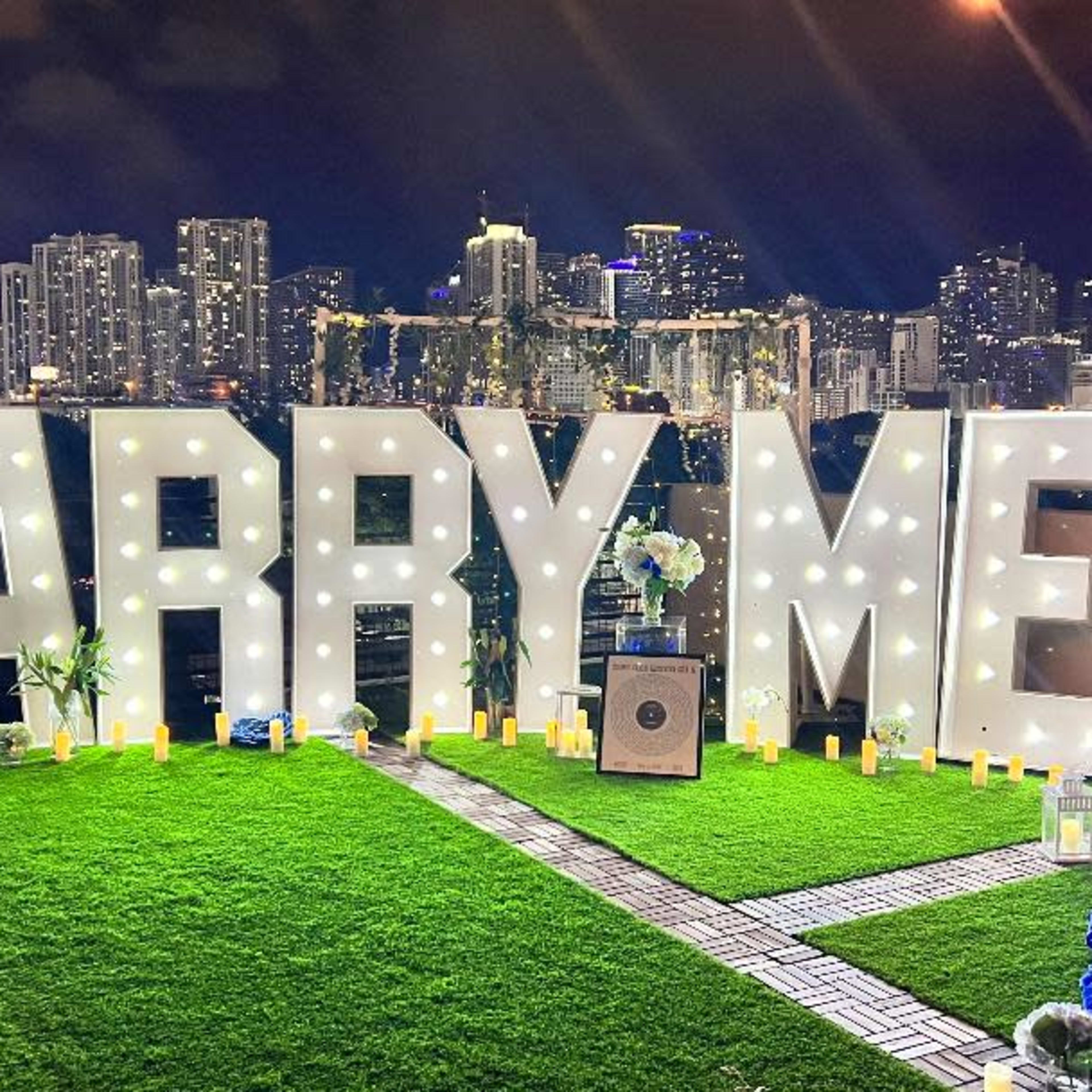 The scene features a large "MARRY ME" display illuminated with lights on a rooftop, surrounded by candles and flowers, with a city skyline in the background.