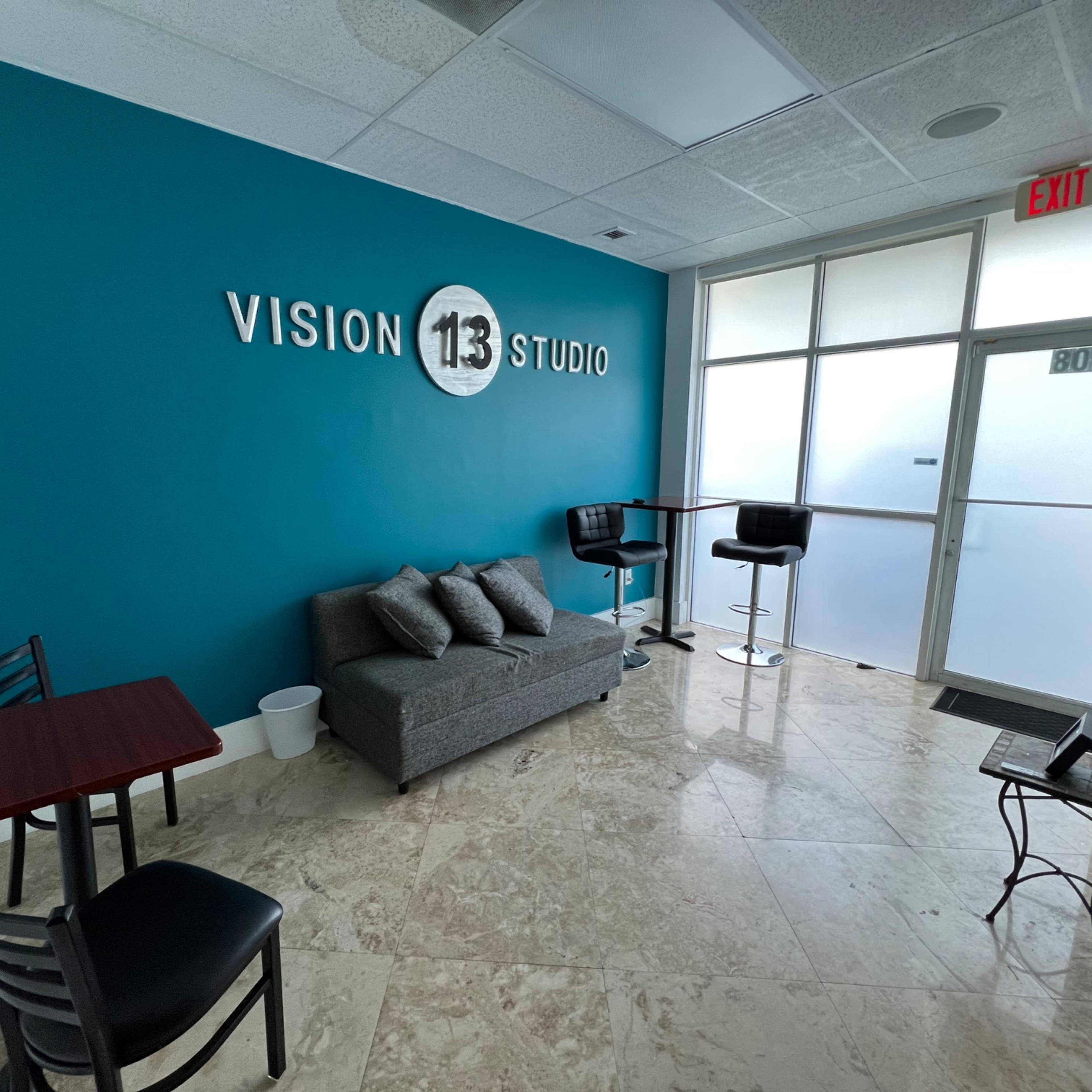 A reception area with a turquoise wall featuring the text "VISION 113 STUDIO," a gray sofa, a small table, and bar stools next to a large glass door.