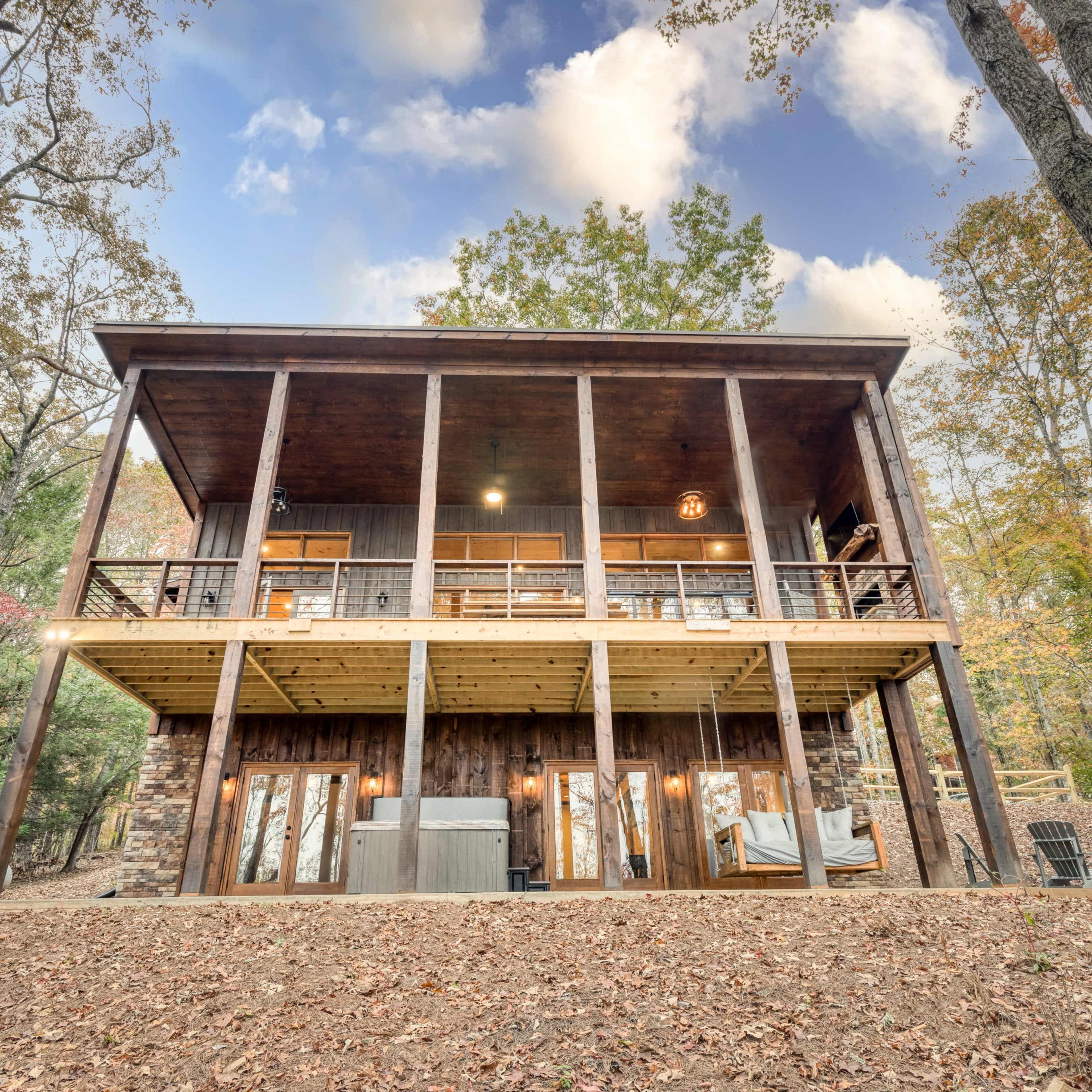 A two-story wooden cabin with a spacious deck overlooks a landscaped area surrounded by trees and fallen leaves.