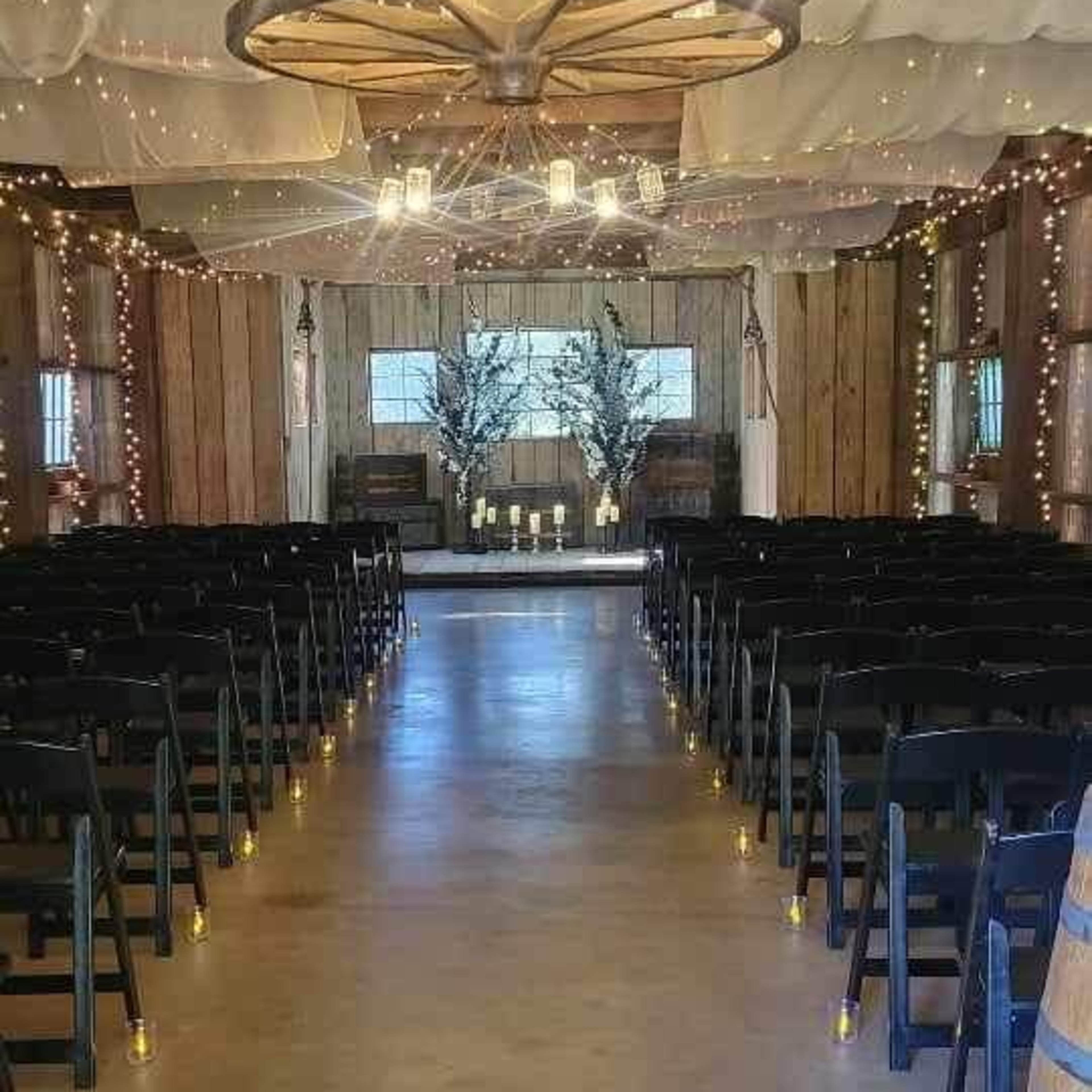 The interior of a rustic venue is set up for a wedding ceremony, featuring rows of black chairs, decorative lighting, and a decorated altar at the far end.