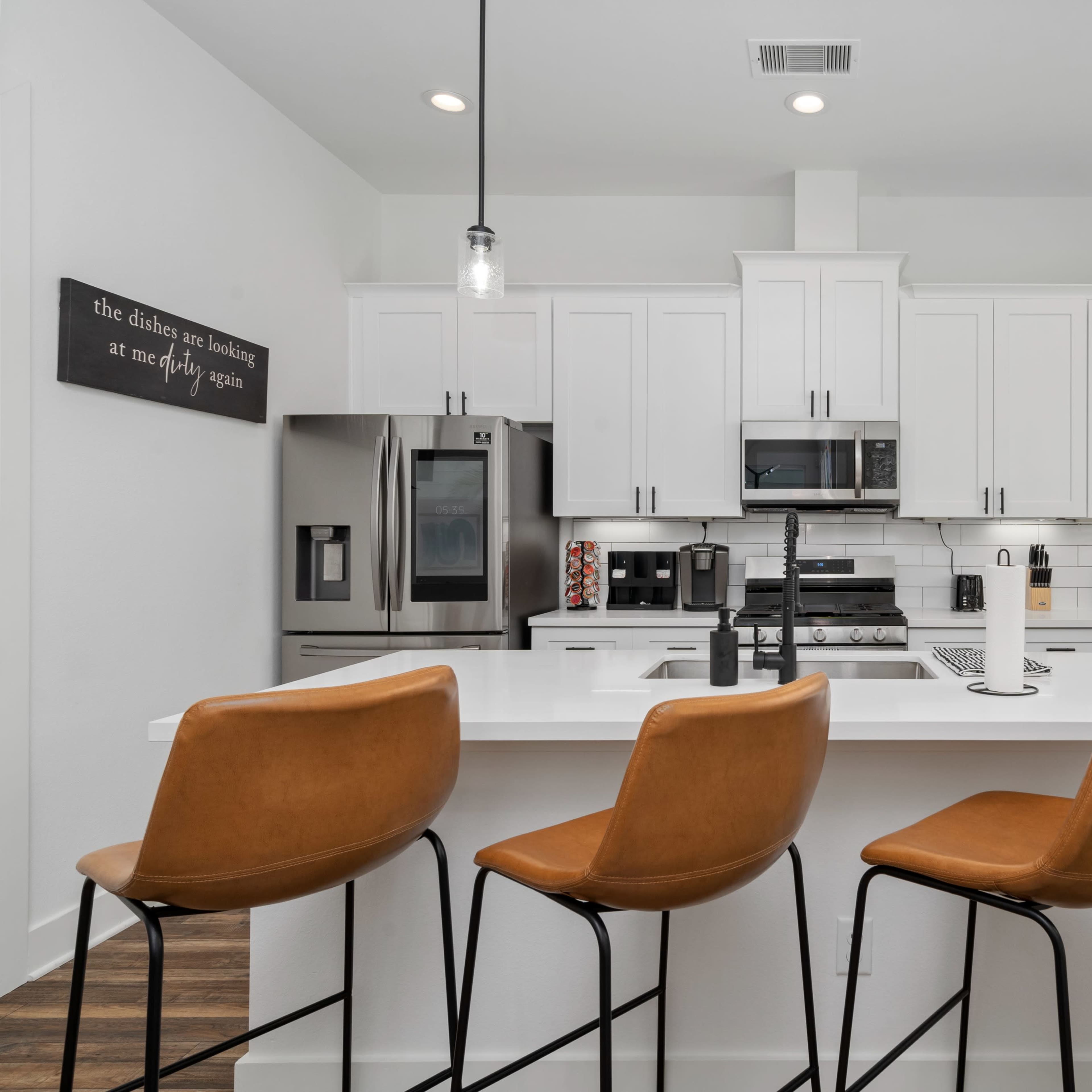 A modern kitchen featuring a white island with four tan bar stools, stainless steel appliances, and bright cabinetry.