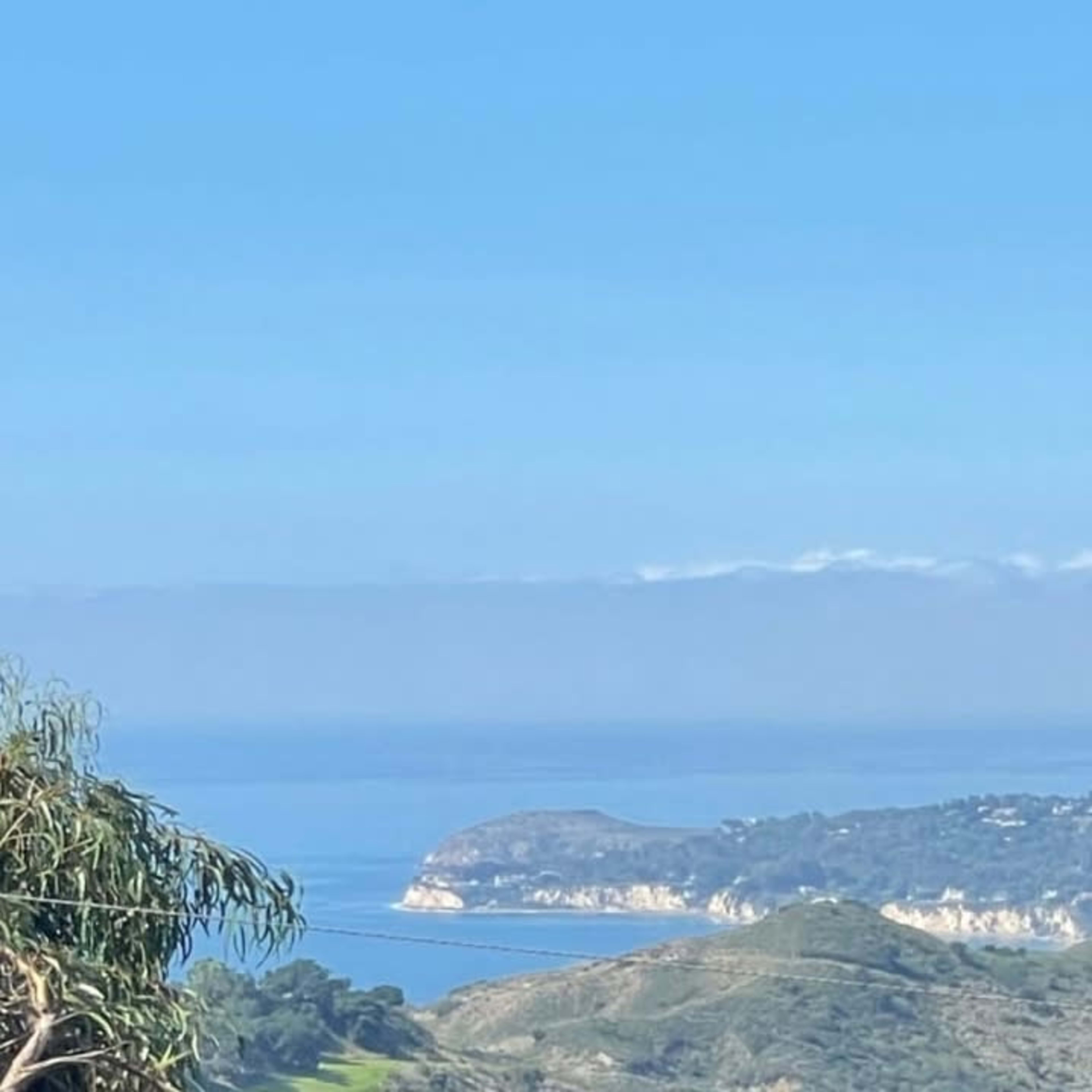 A panoramic view of a coastal landscape featuring hills, ocean, and distant cliffs under a clear blue sky.