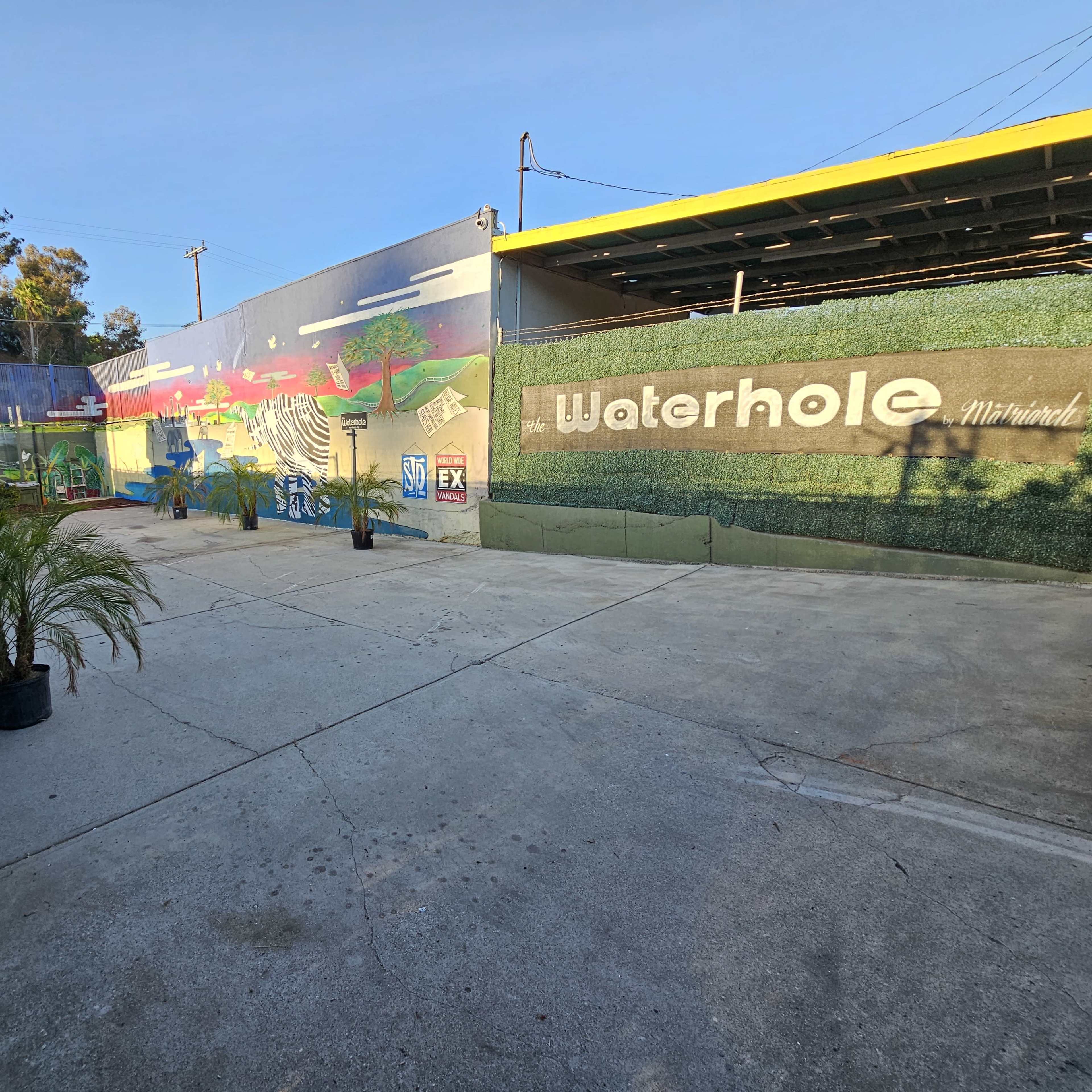 A spacious outdoor area featuring a mural on a wall labeled "Waterhole," with concrete flooring and potted plants.