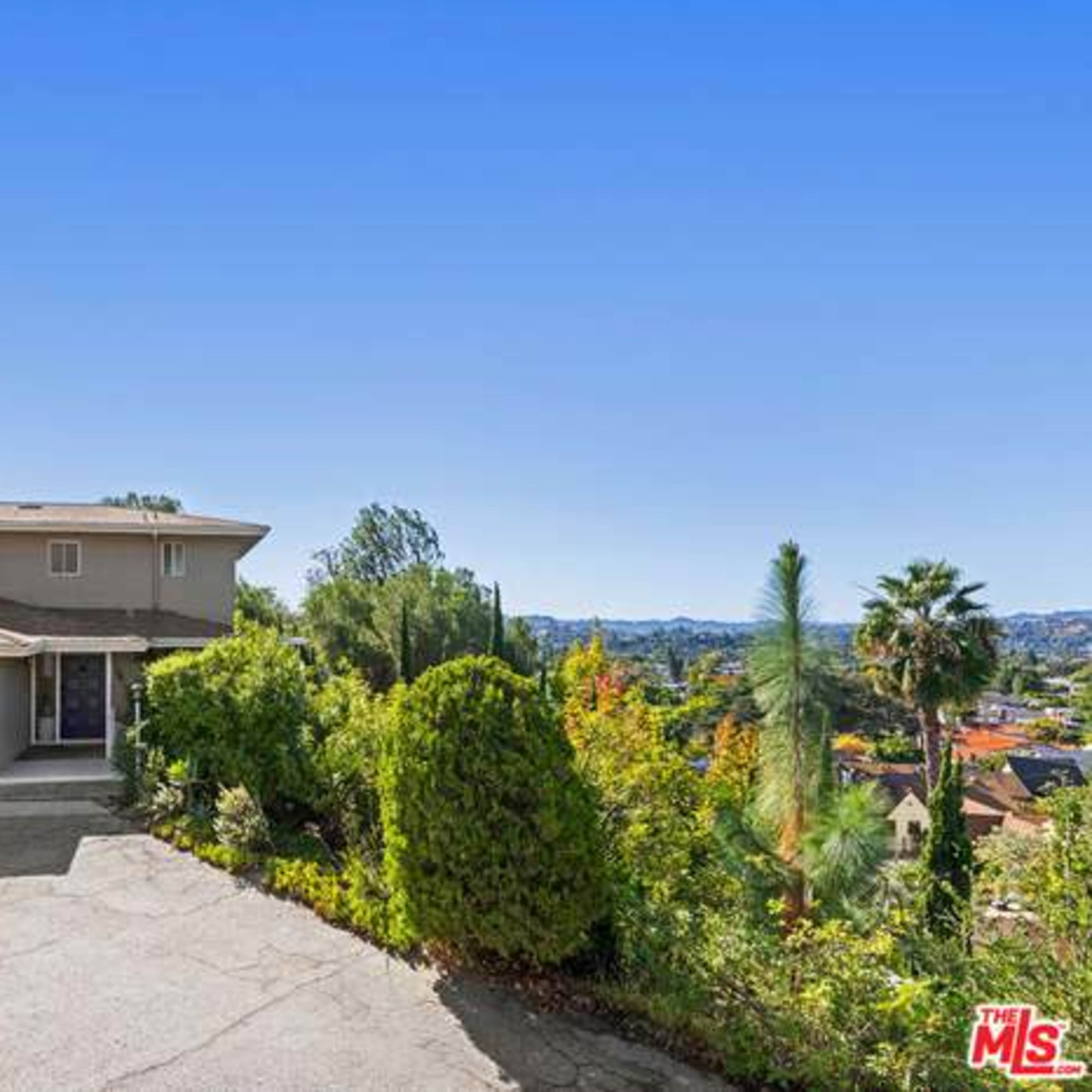 A two-story house with a driveway leading up to a garage, surrounded by landscaped greenery and distant hills.