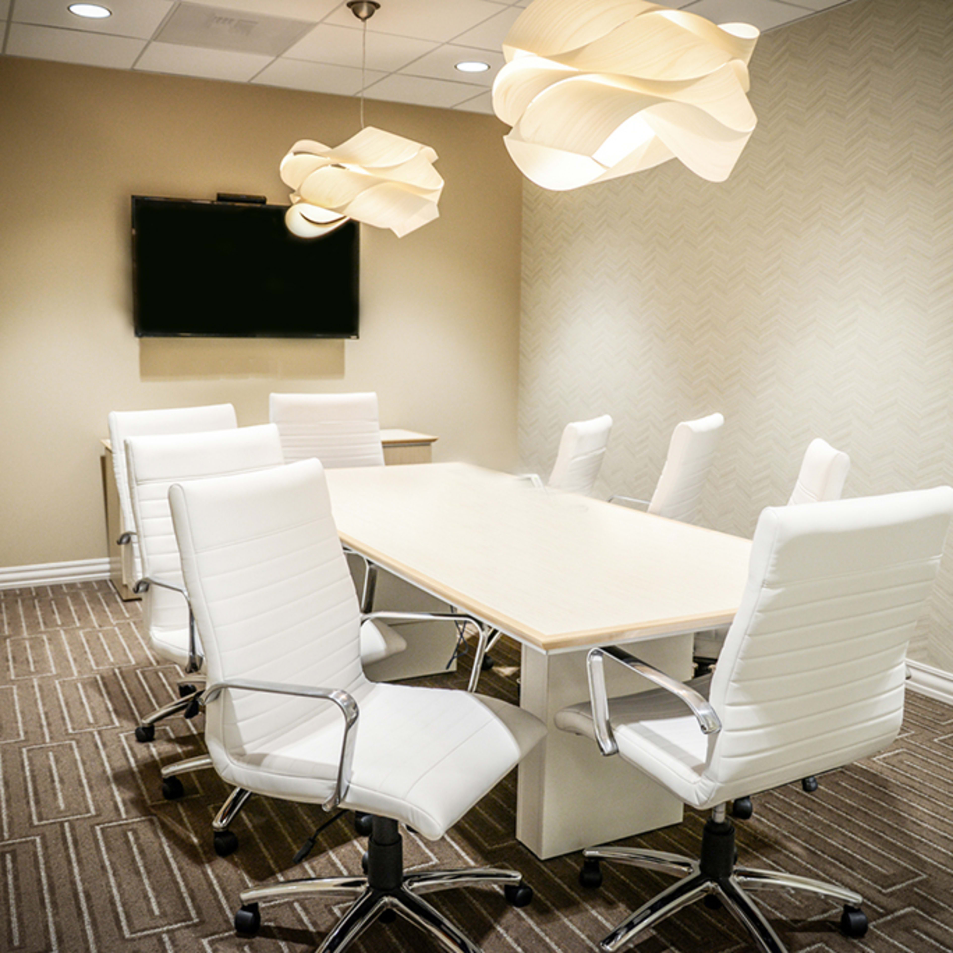 A modern conference room with a large table surrounded by six white ergonomic chairs and two pendant lights hanging from the ceiling.