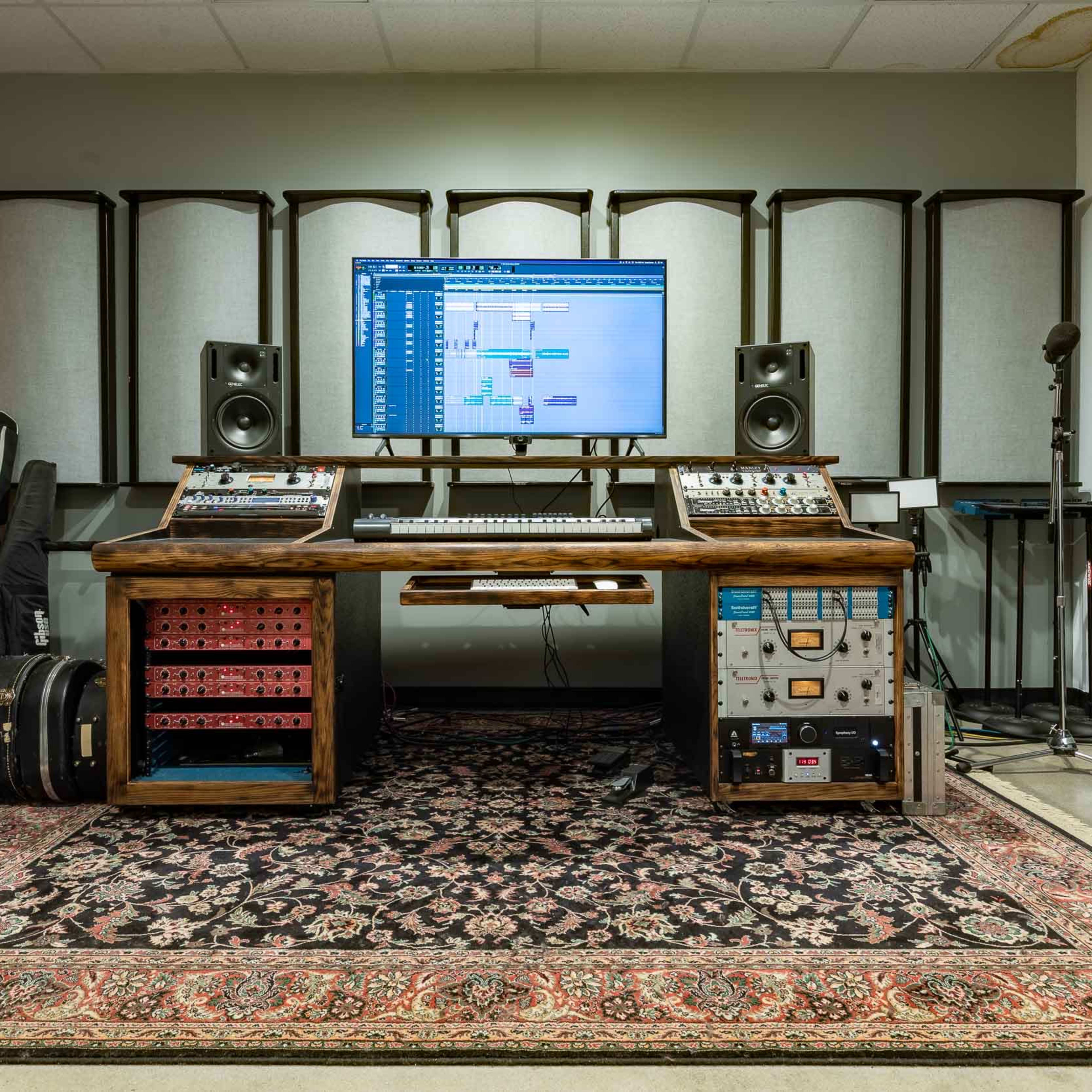 A music production studio features a large wooden desk with audio equipment, a computer display, and surrounding soundproof panels.