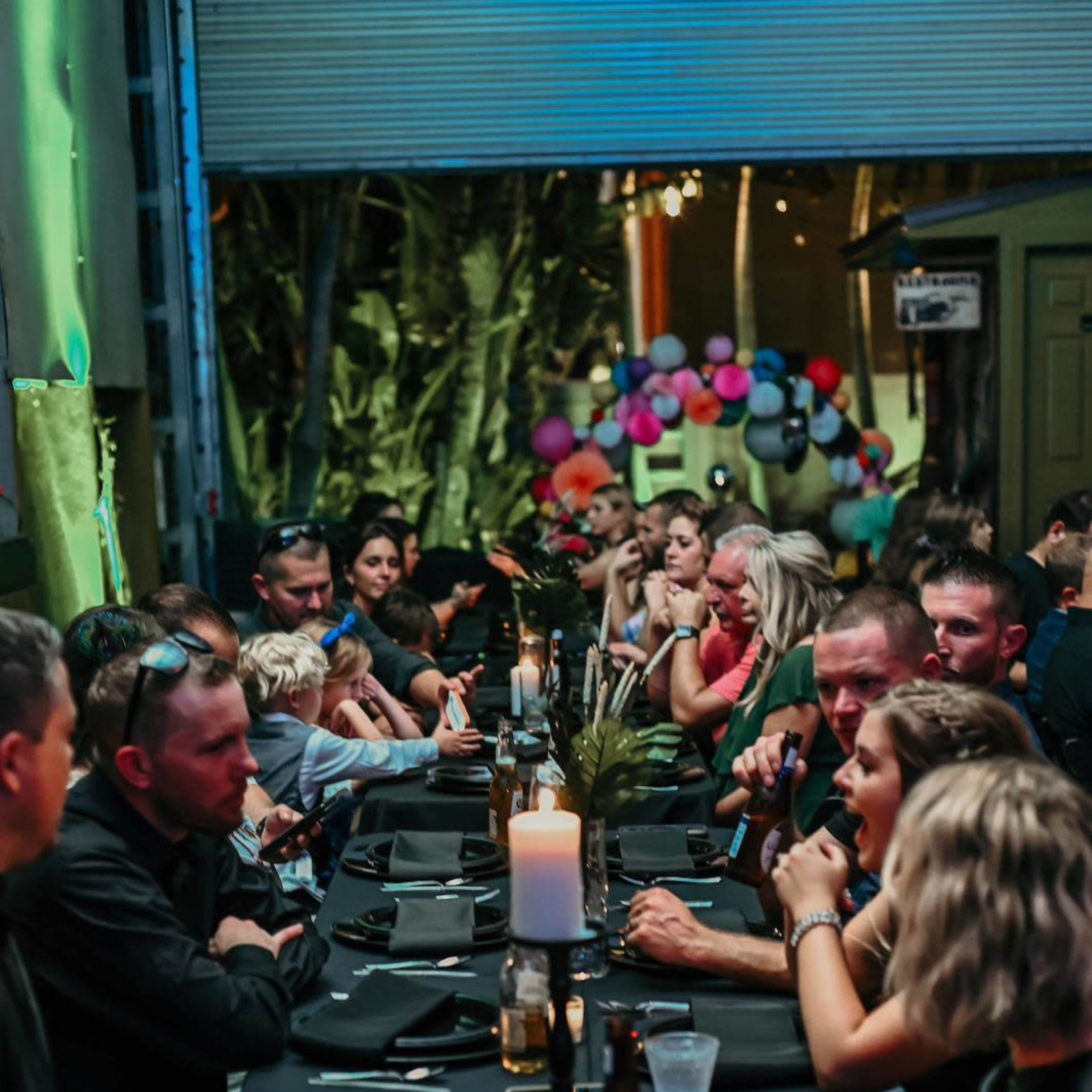A long table with guests seated on both sides is set for a gathering, featuring drinks and a candle in the center, while colorful decorations are visible in the background.