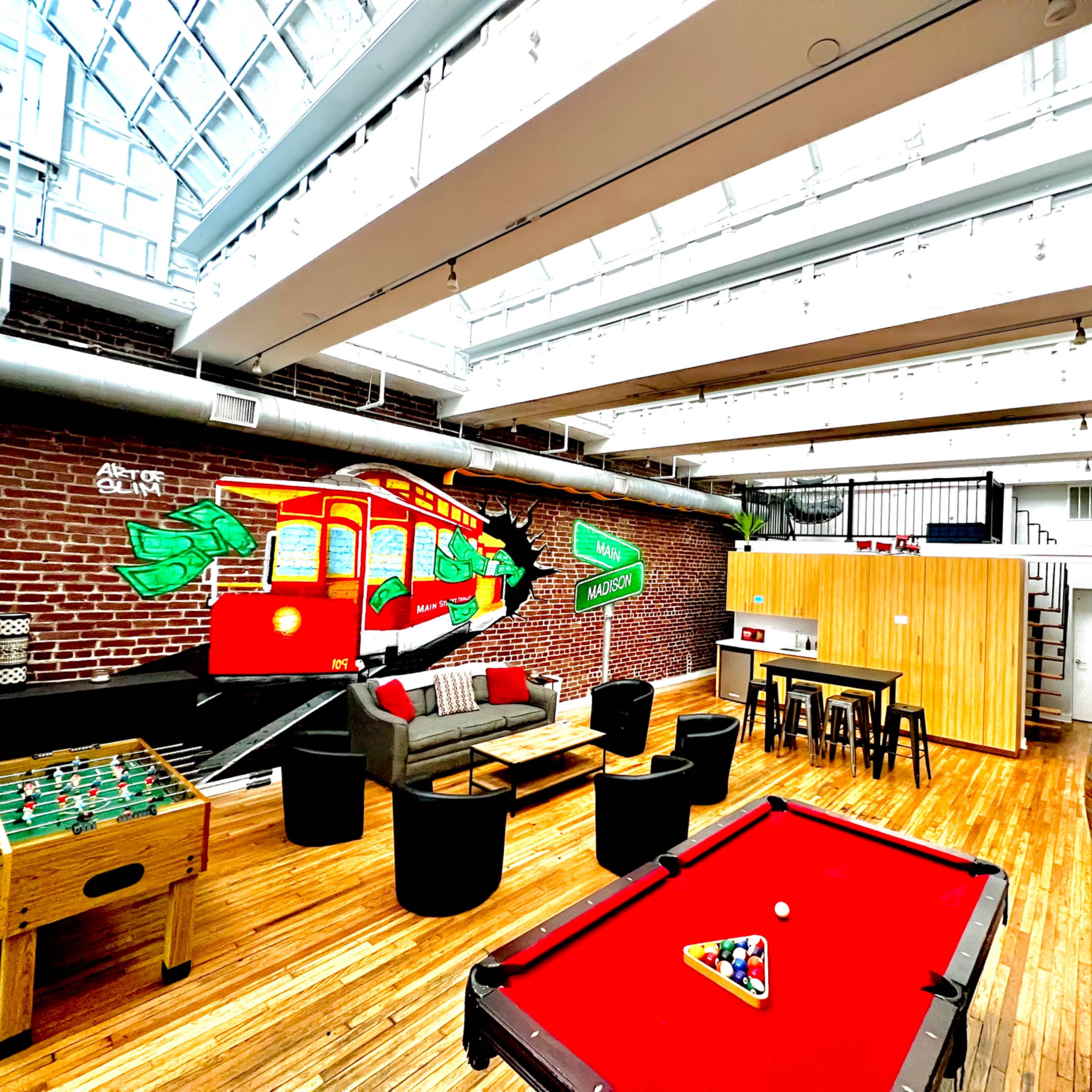 A spacious room with a pool table, foosball table, and a mural of a trolley on a brick wall, all under a glass ceiling.