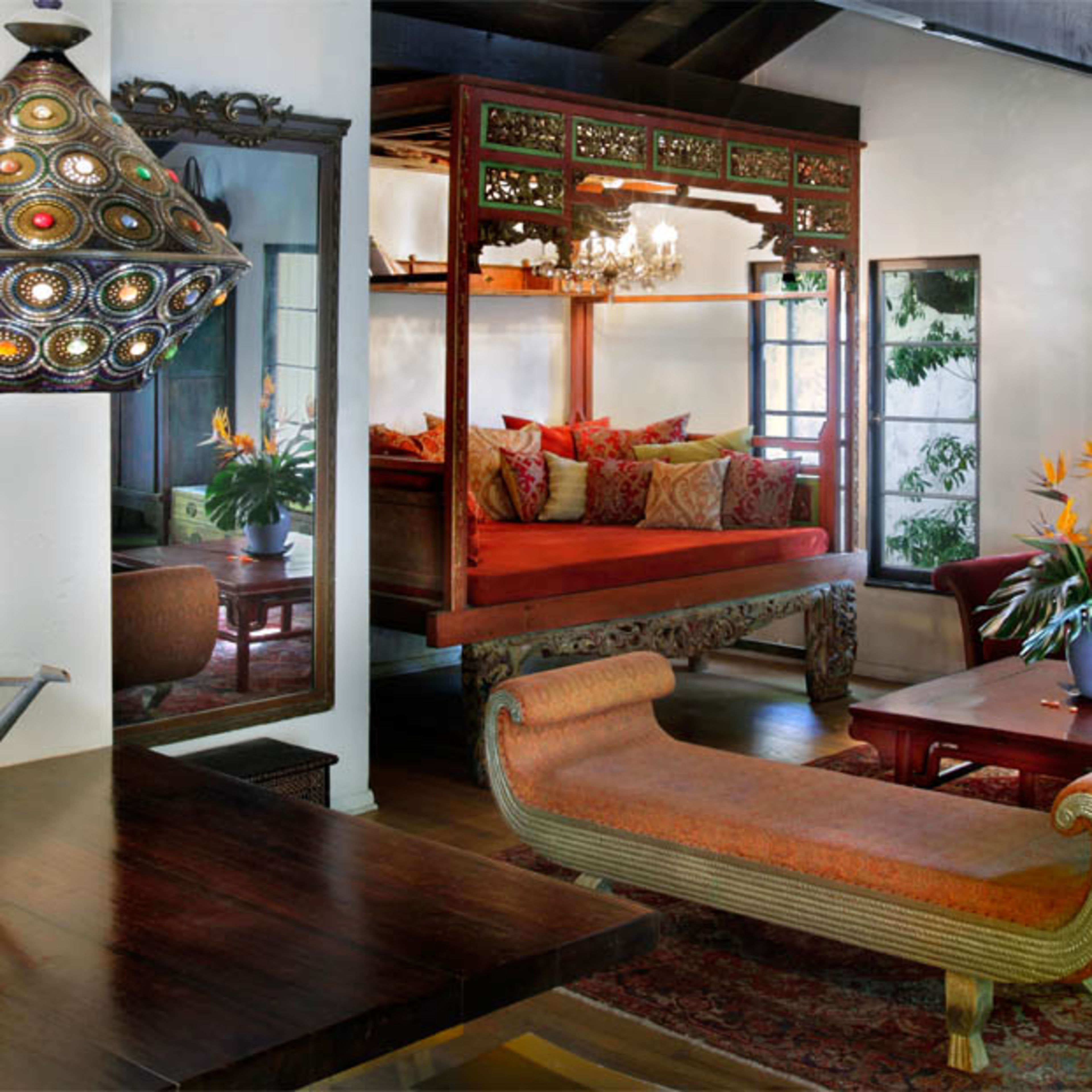 A brightly decorated living room features a large, ornate bed, a wooden table with floral arrangements, and various pieces of art on the walls.