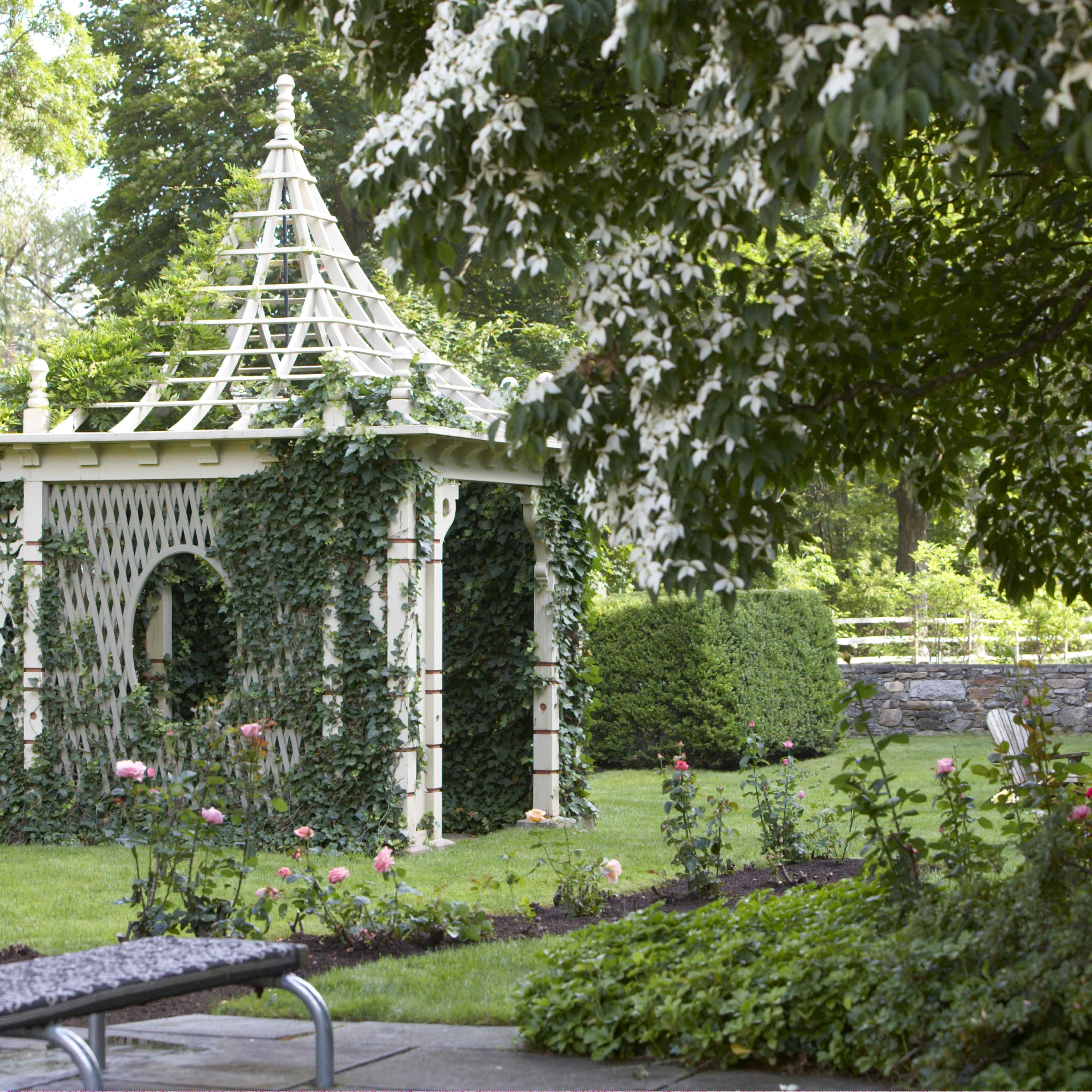 A white gazebo covered in ivy stands surrounded by a well-maintained garden with blooming flowers and lush greenery.