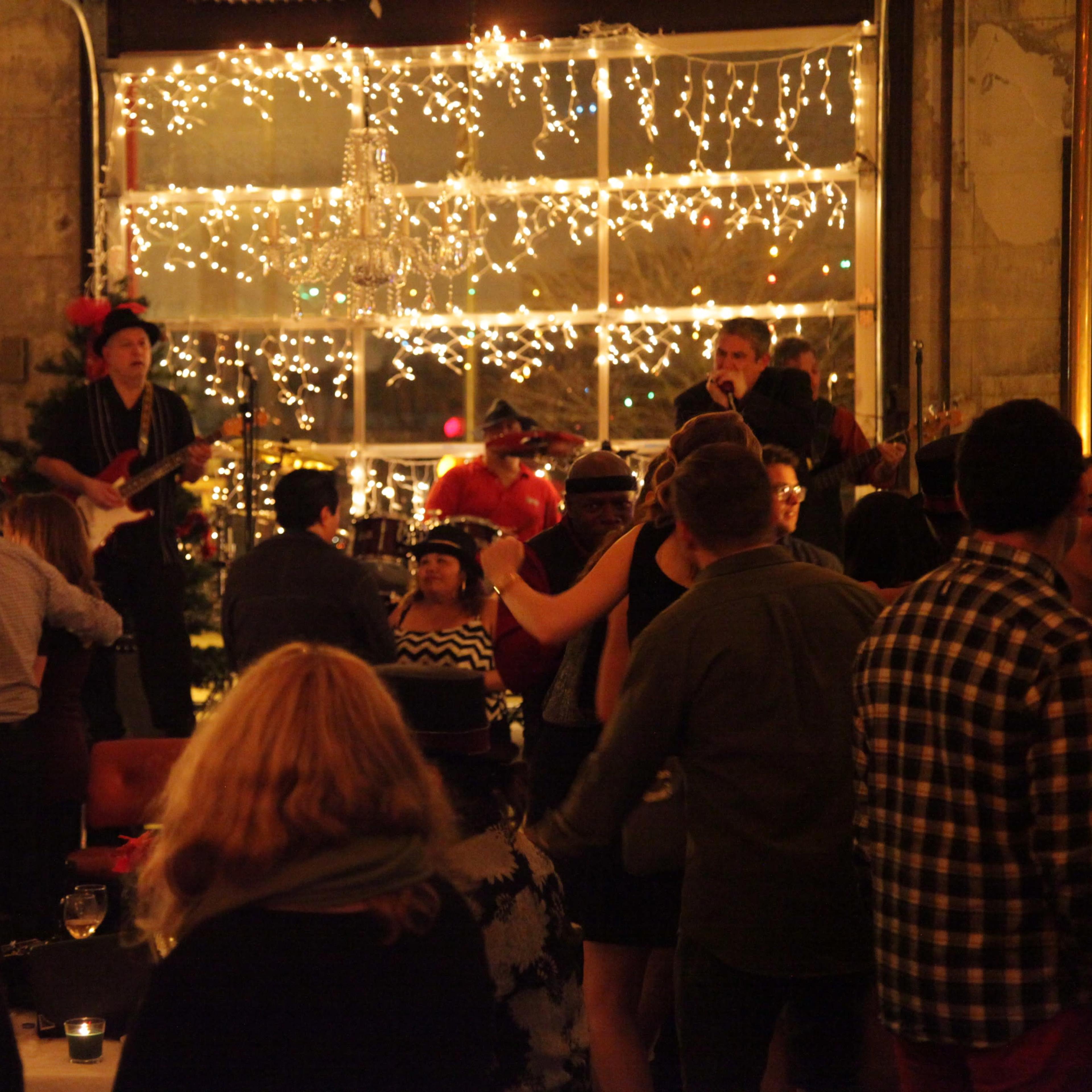 The scene depicts a lively indoor party with guests dancing and a band playing, illuminated by string lights and a chandelier.