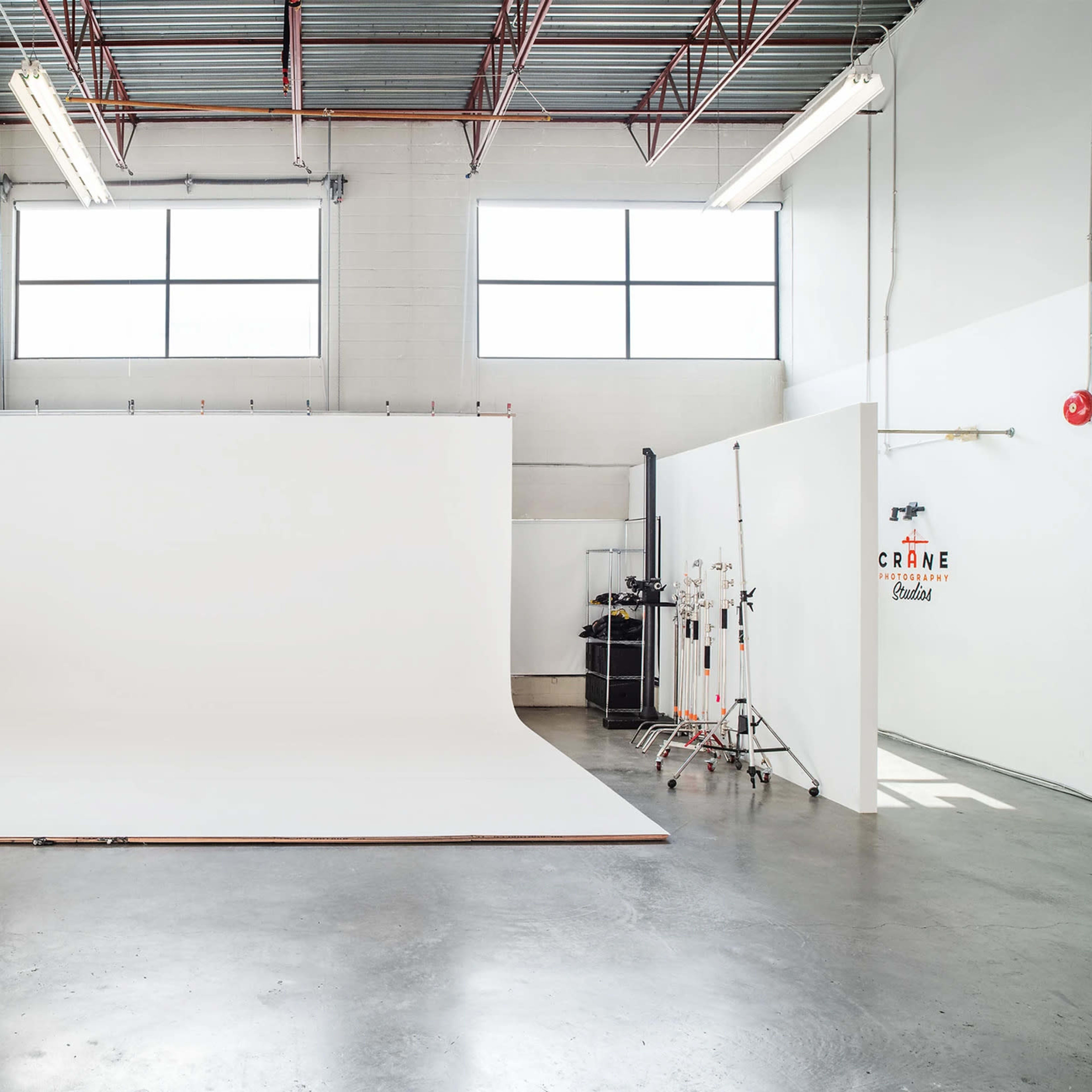 An empty photo studio features a large white backdrop, bright natural light from windows, and gray concrete floors.