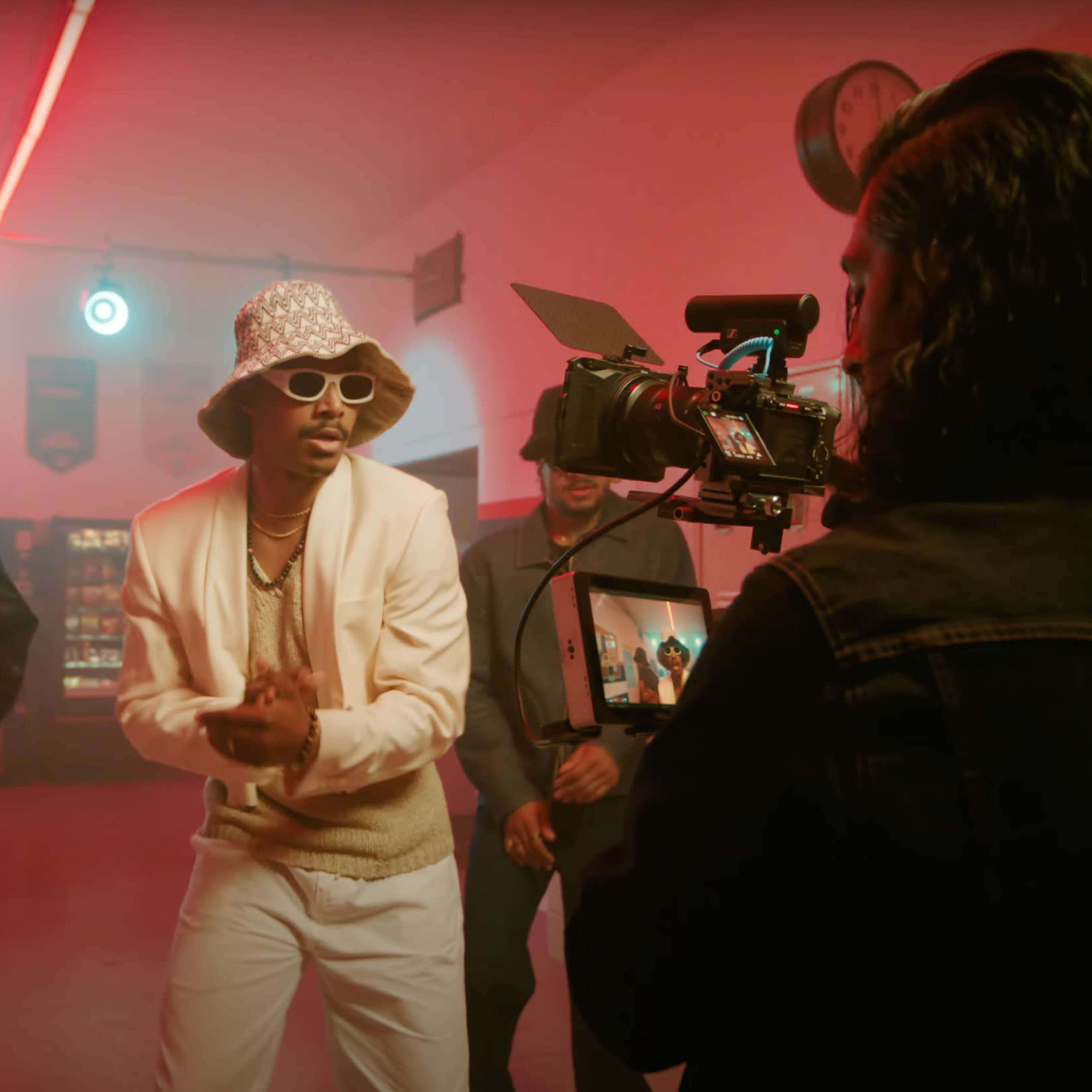 A music video shoot takes place in a brightly lit hallway, where a performer dances while being filmed by a crew member holding a camera.
