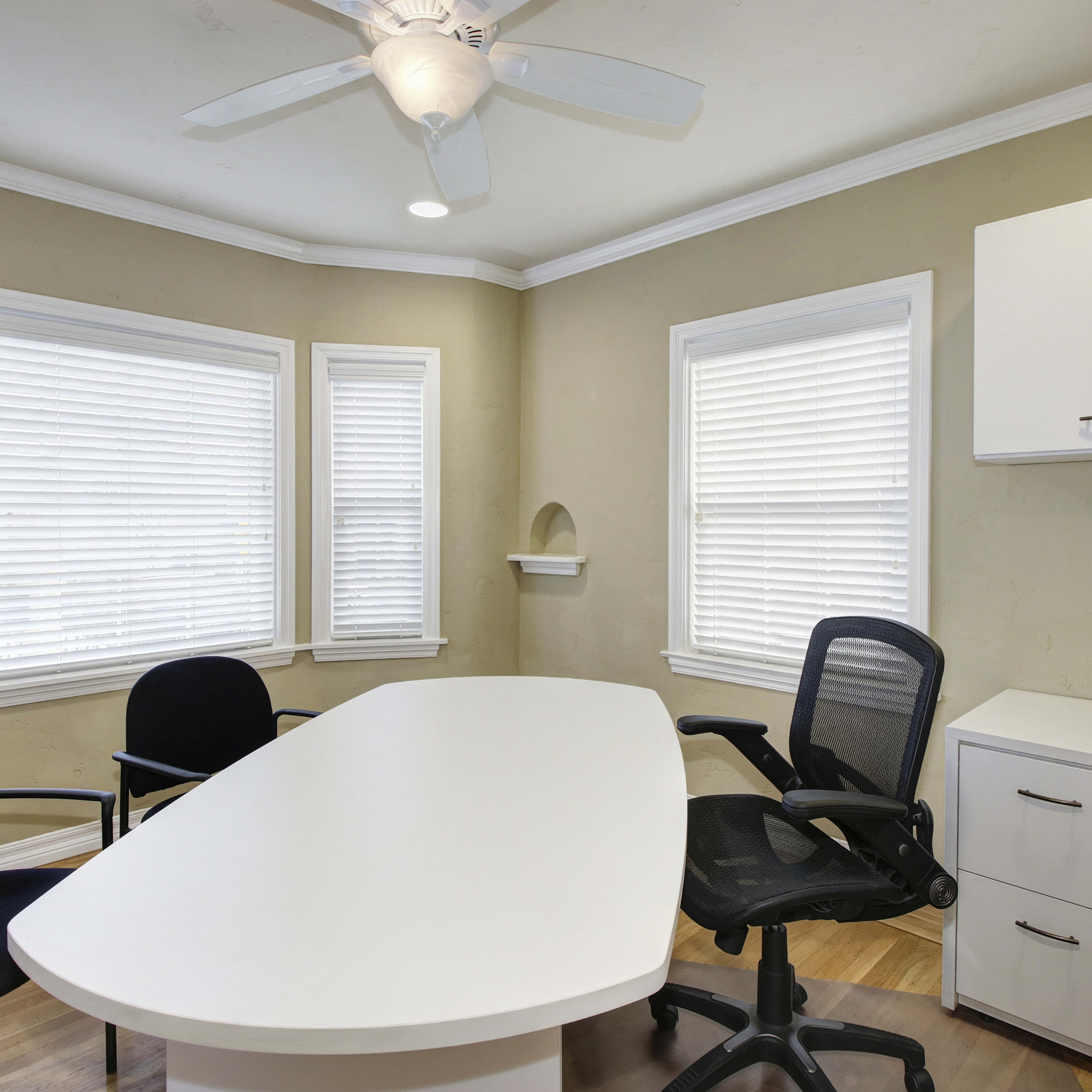 A modern office space features a white conference table, two black chairs, a desk with storage, and multiple windows with blinds.