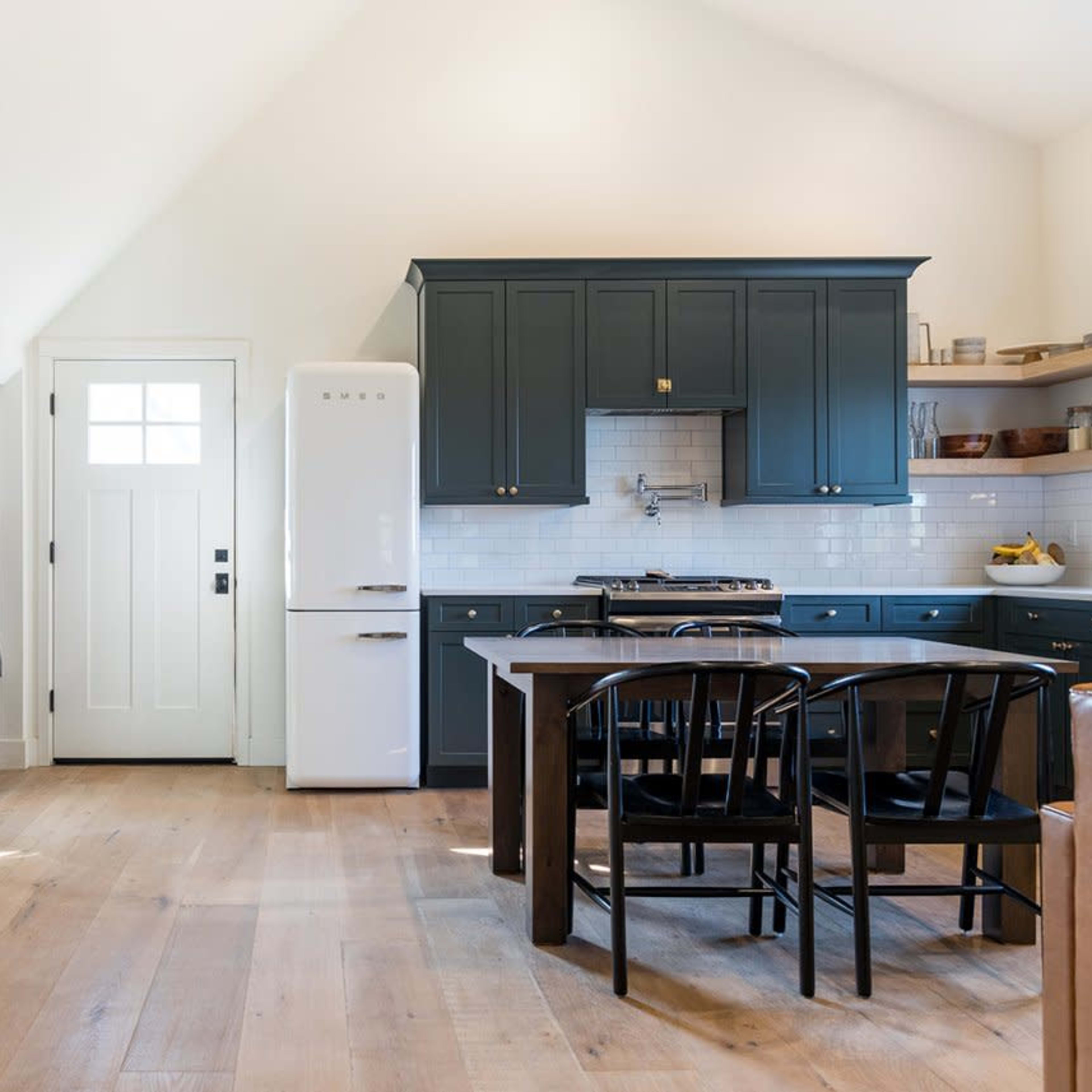 A modern kitchen features a dark blue cabinetry, a white refrigerator, and a wooden dining table with black chairs.