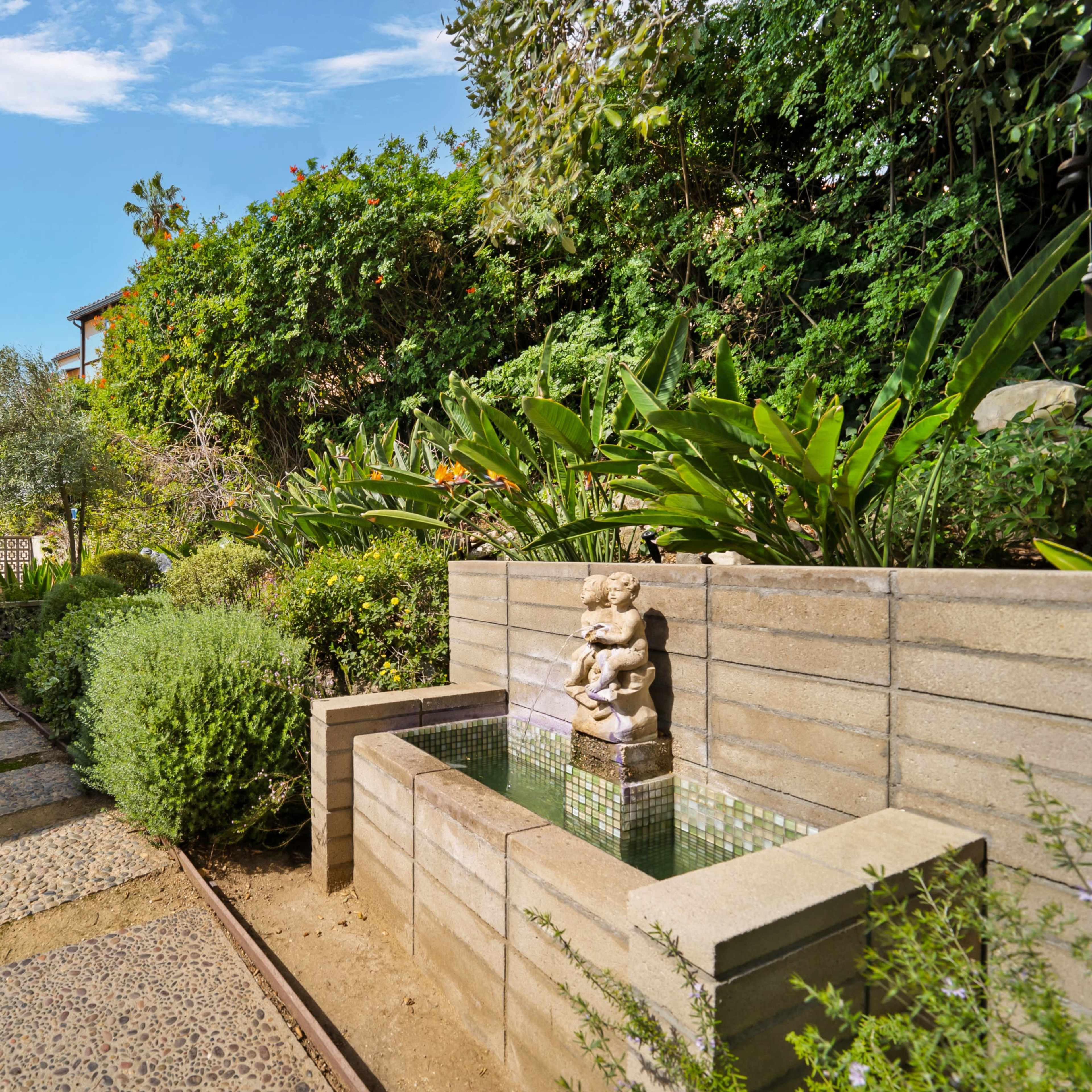 A stone pathway leads to a landscaped garden featuring a wall-mounted fountain with a statue.