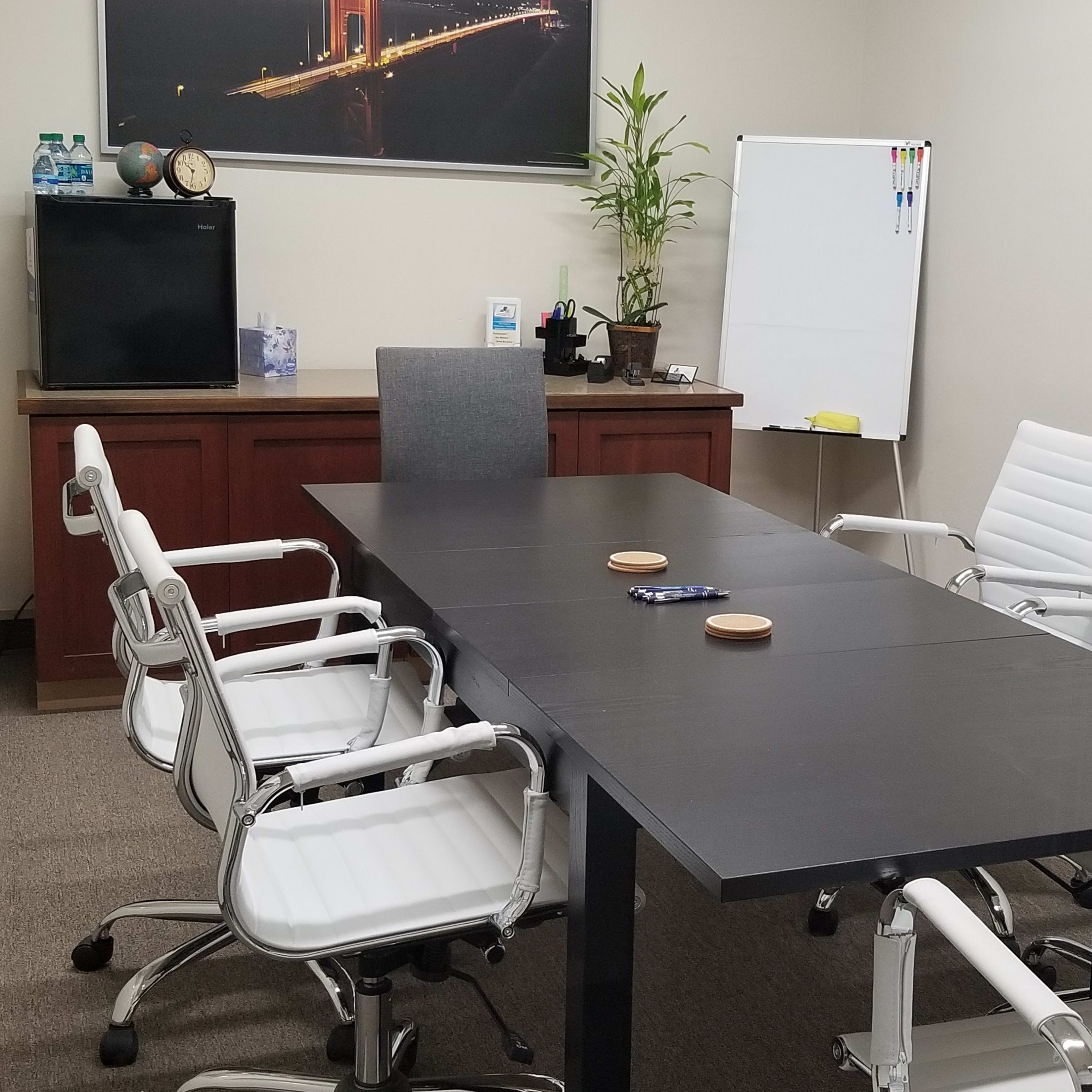A modern conference room features a black table surrounded by white chairs, with a wall-mounted picture, a plant, and a whiteboard in the background.