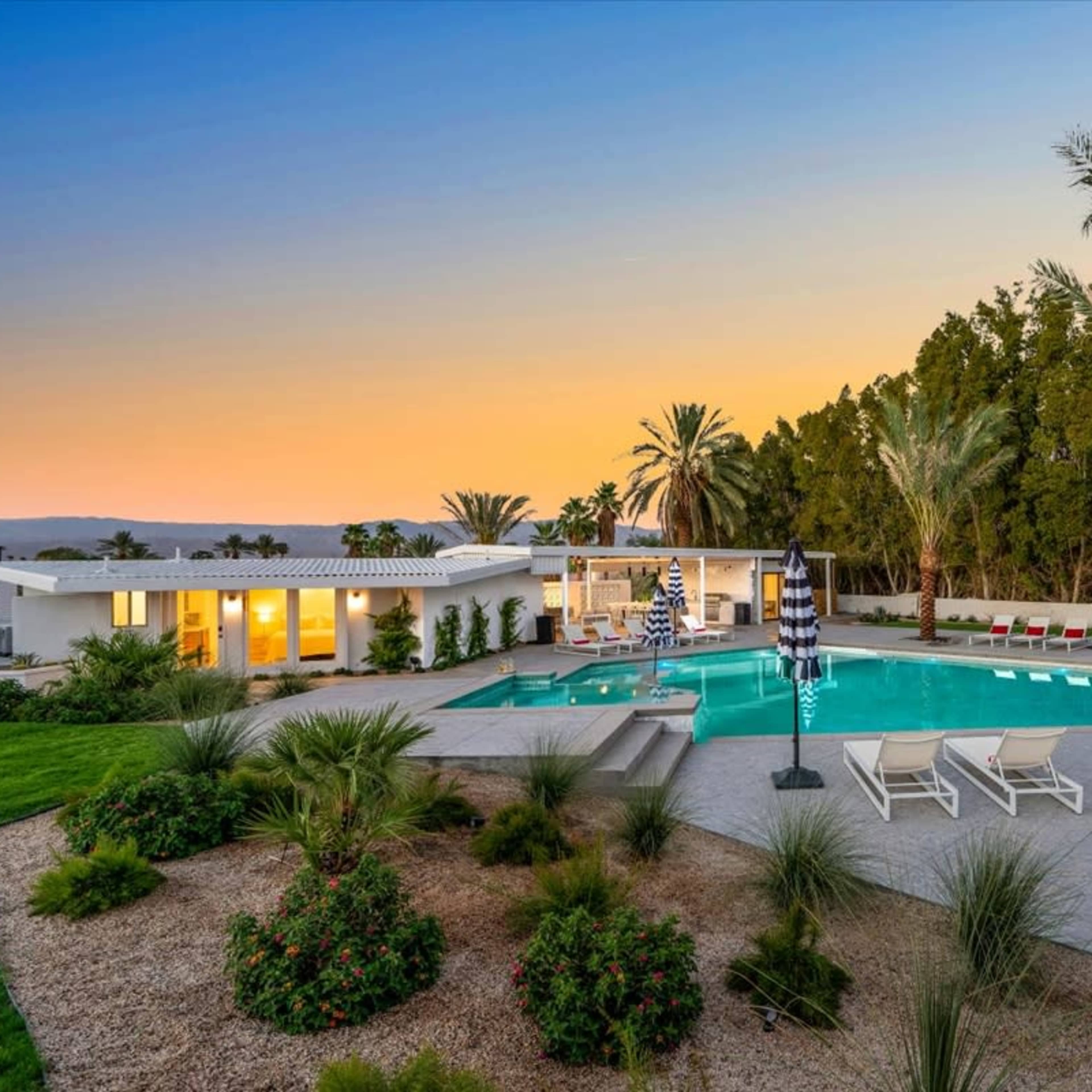 A modern home with a swimming pool surrounded by lounge chairs and palm trees, set against a sunset sky.