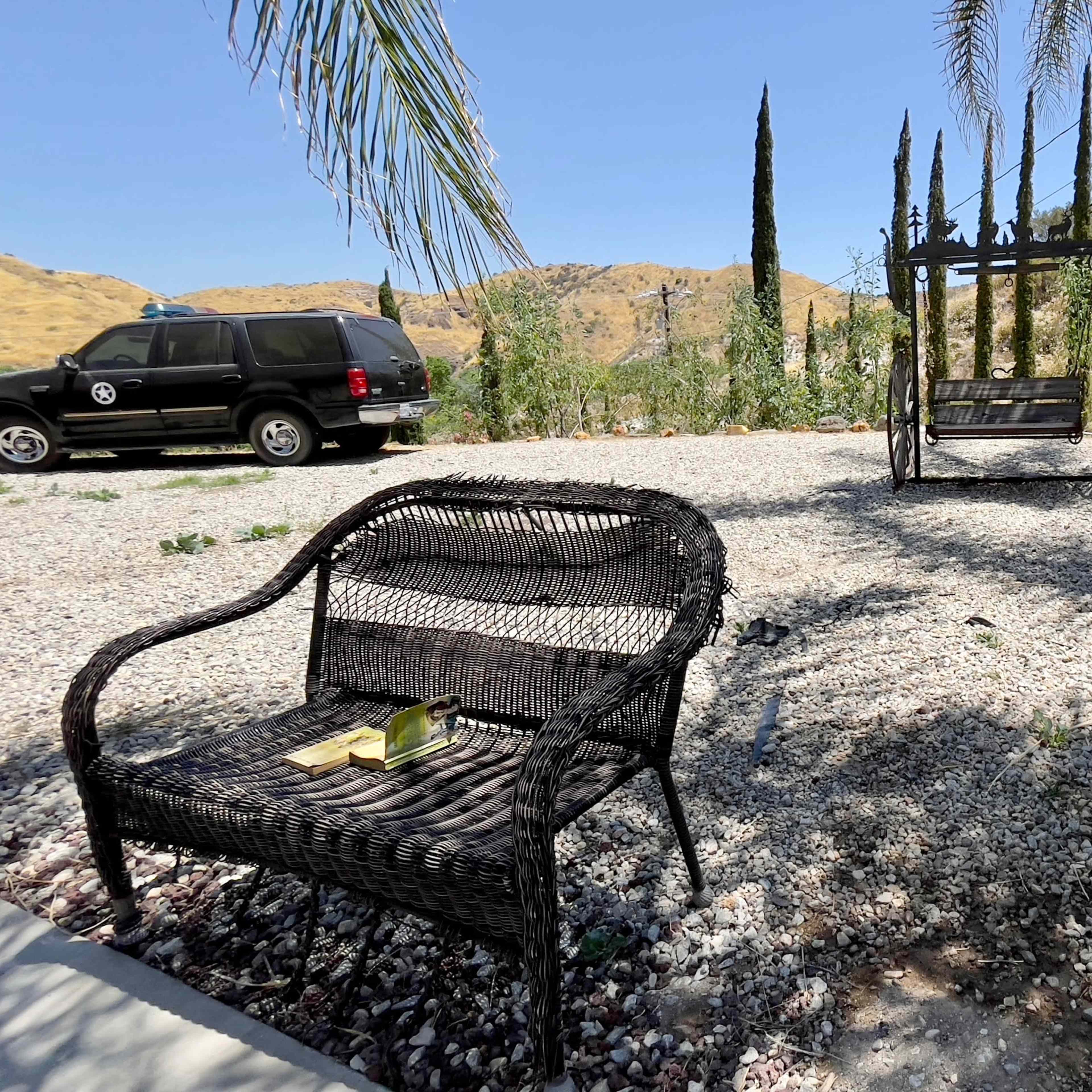 A black SUV is parked near a gravel area with a wicker chair and a swing surrounded by tall trees and hills.
