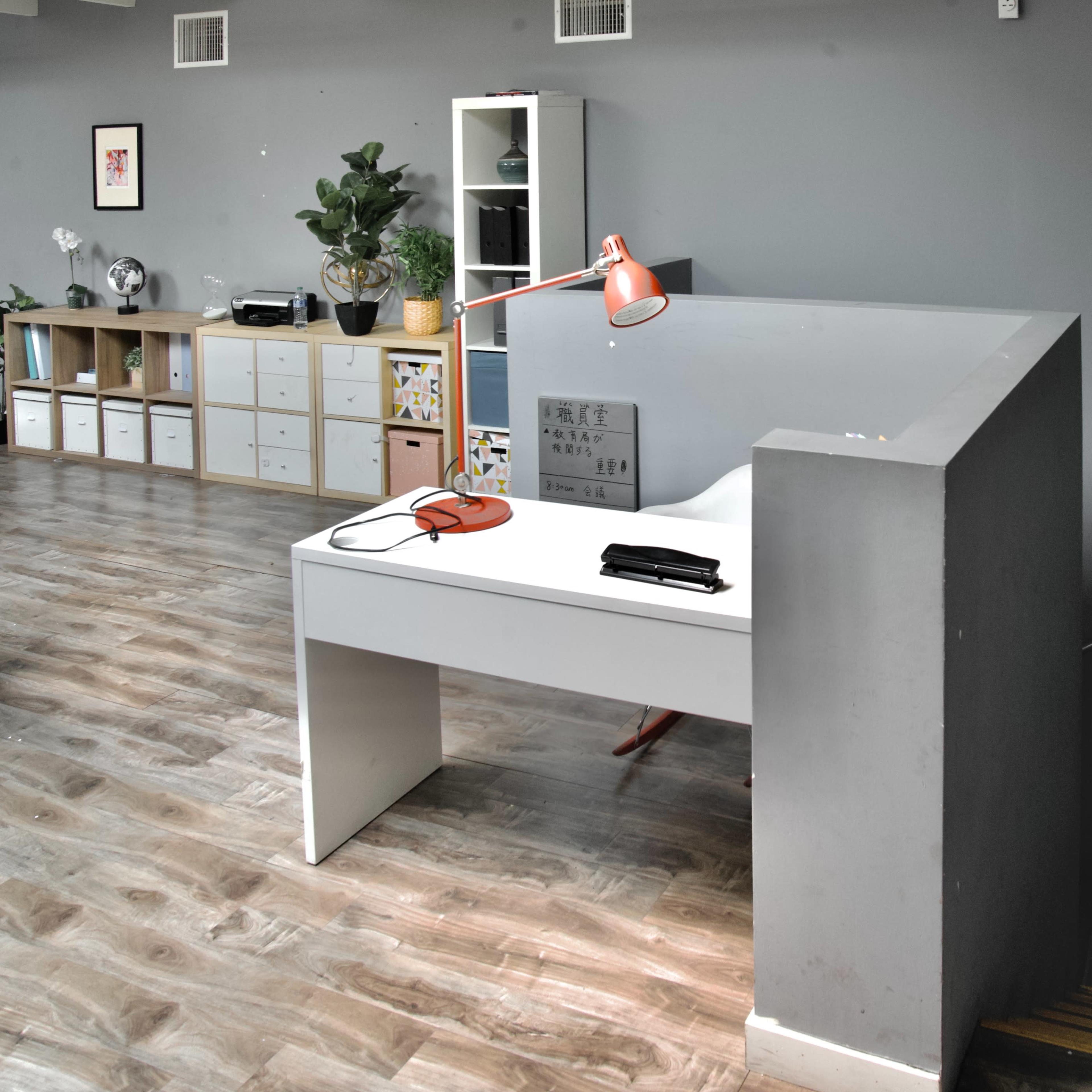 A modern office space with a white desk, a red desk lamp, a chair, and storage units against a gray wall.