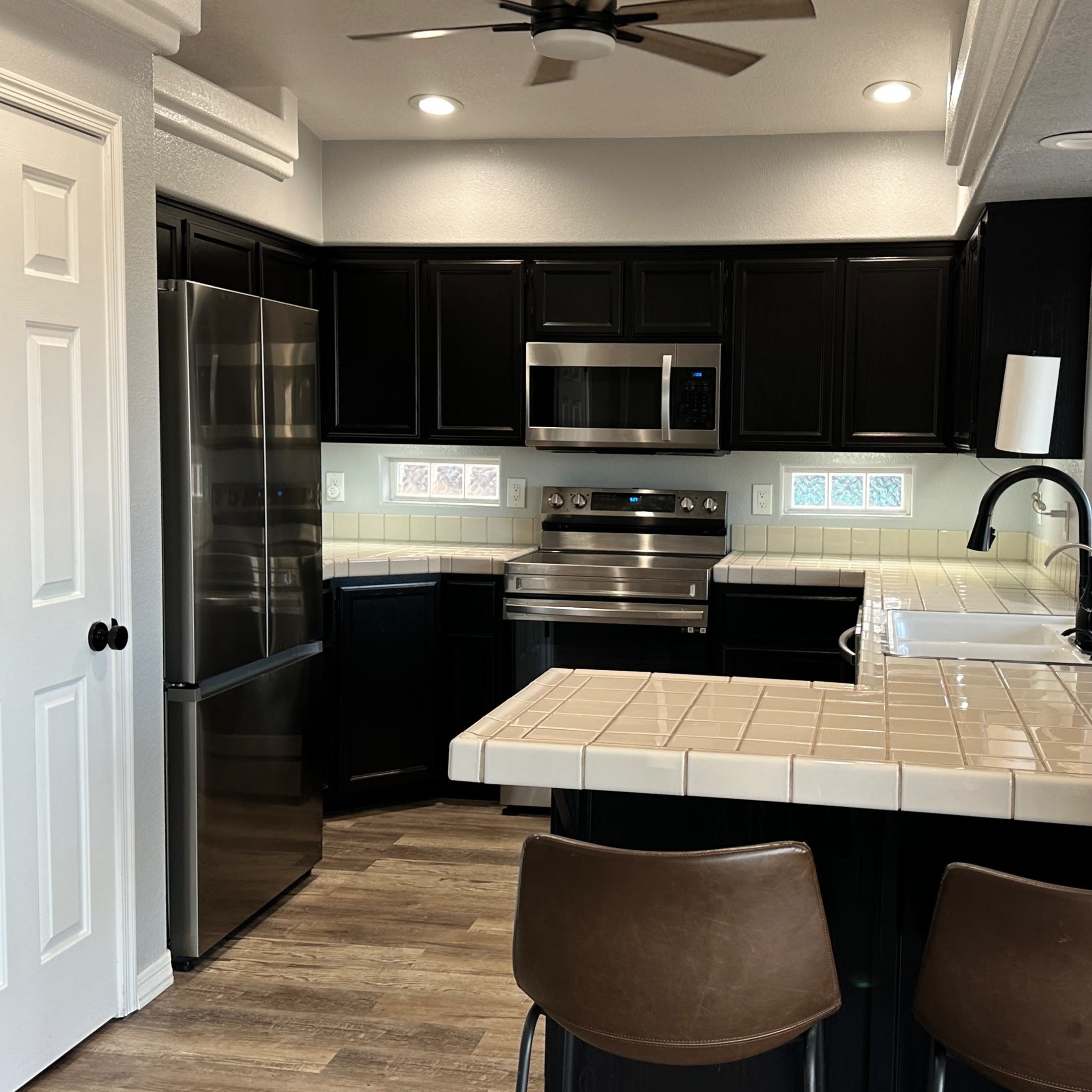 A modern kitchen with black cabinetry, stainless steel appliances, and a tiled countertop with two bar stools.