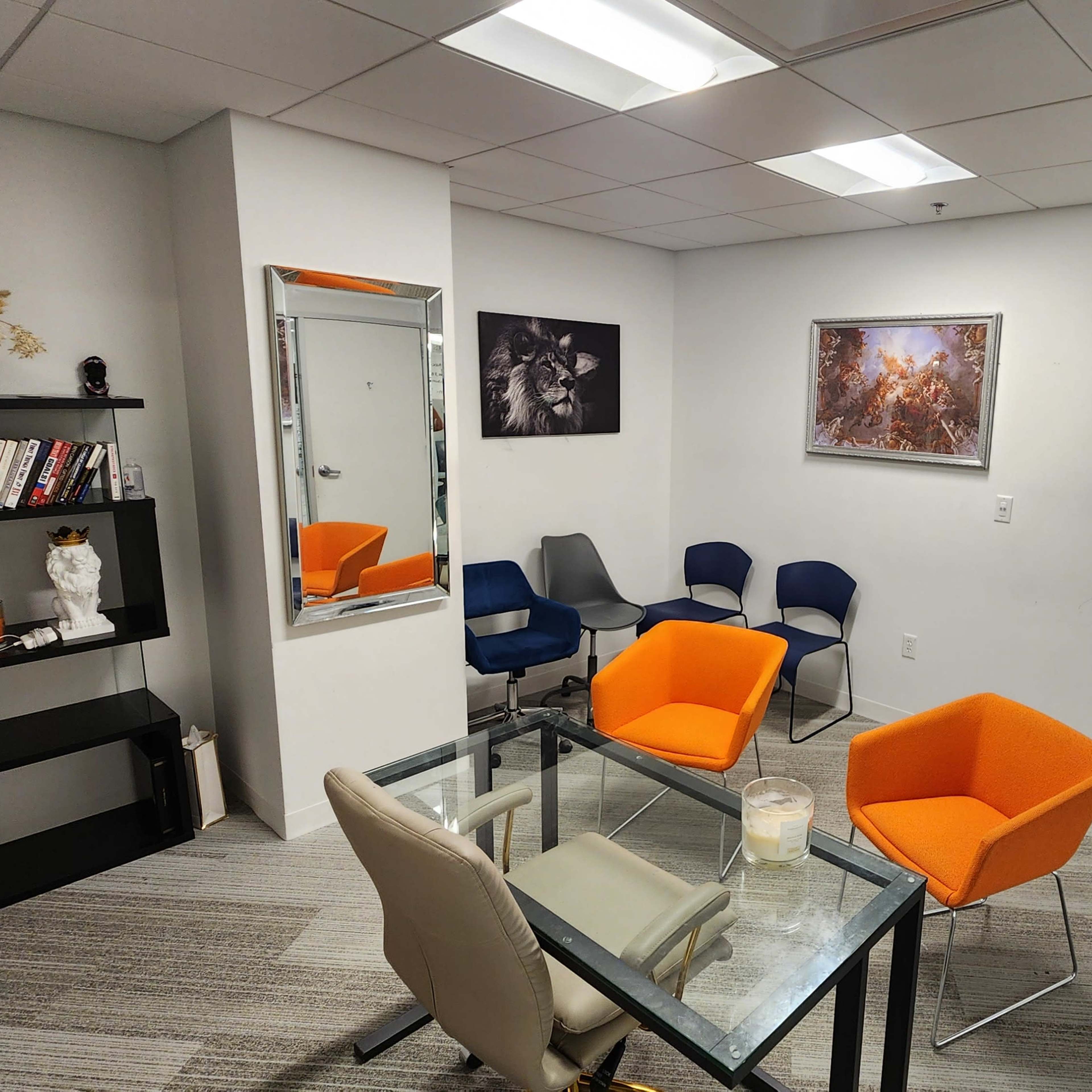 A small meeting room with orange and blue chairs arranged around a glass table, a bookshelf, a mirror, and artwork on the walls.