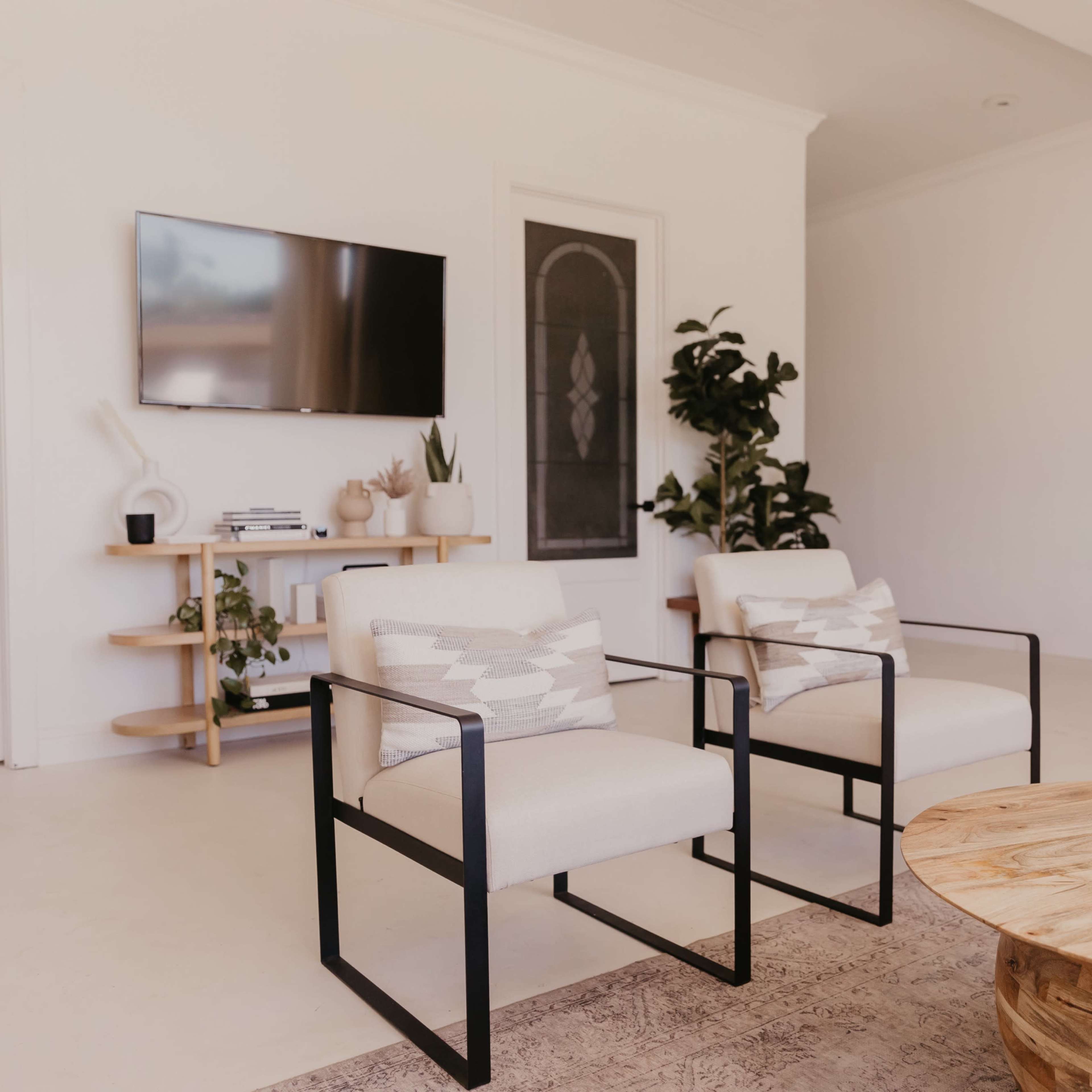 A modern living room with two white armchairs, a round wooden coffee table, a television mounted on the wall, and a corner plant beside a green backdrop.
