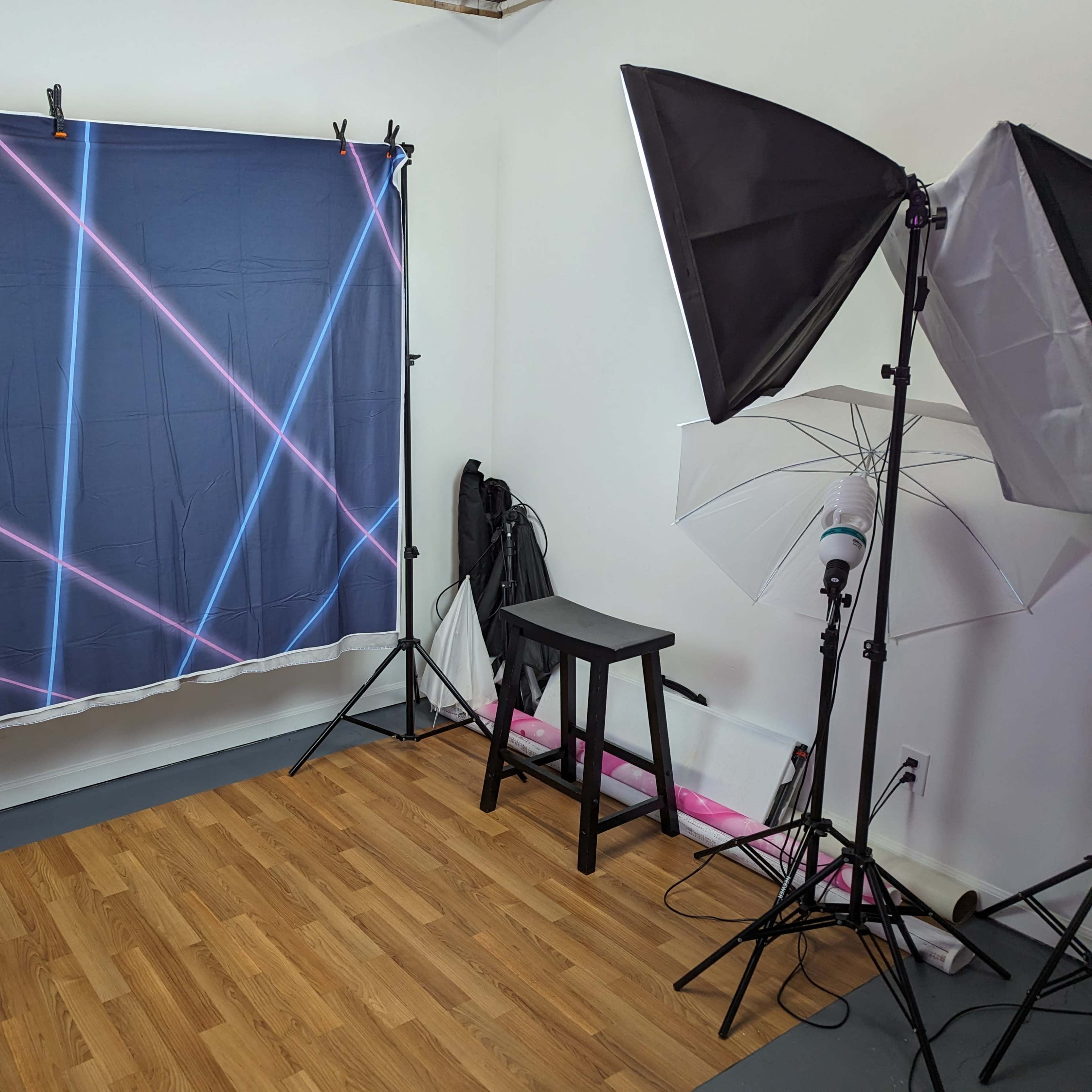 A photography studio setup with a backdrop featuring diagonal neon lines and various lighting equipment, including softboxes and a stool.