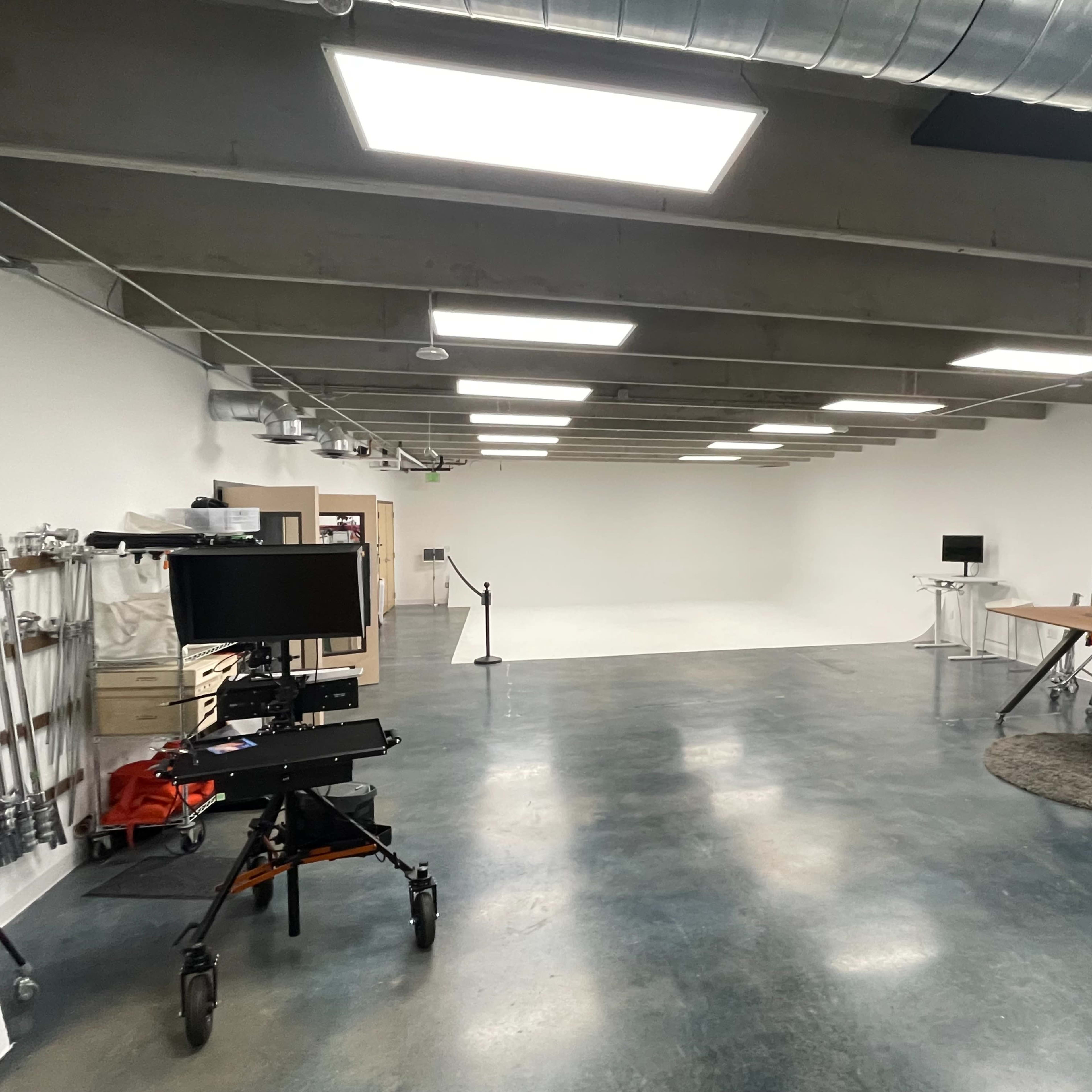 A spacious studio with concrete floors, overhead lighting, and minimal furniture including a large table and equipment on wheels.