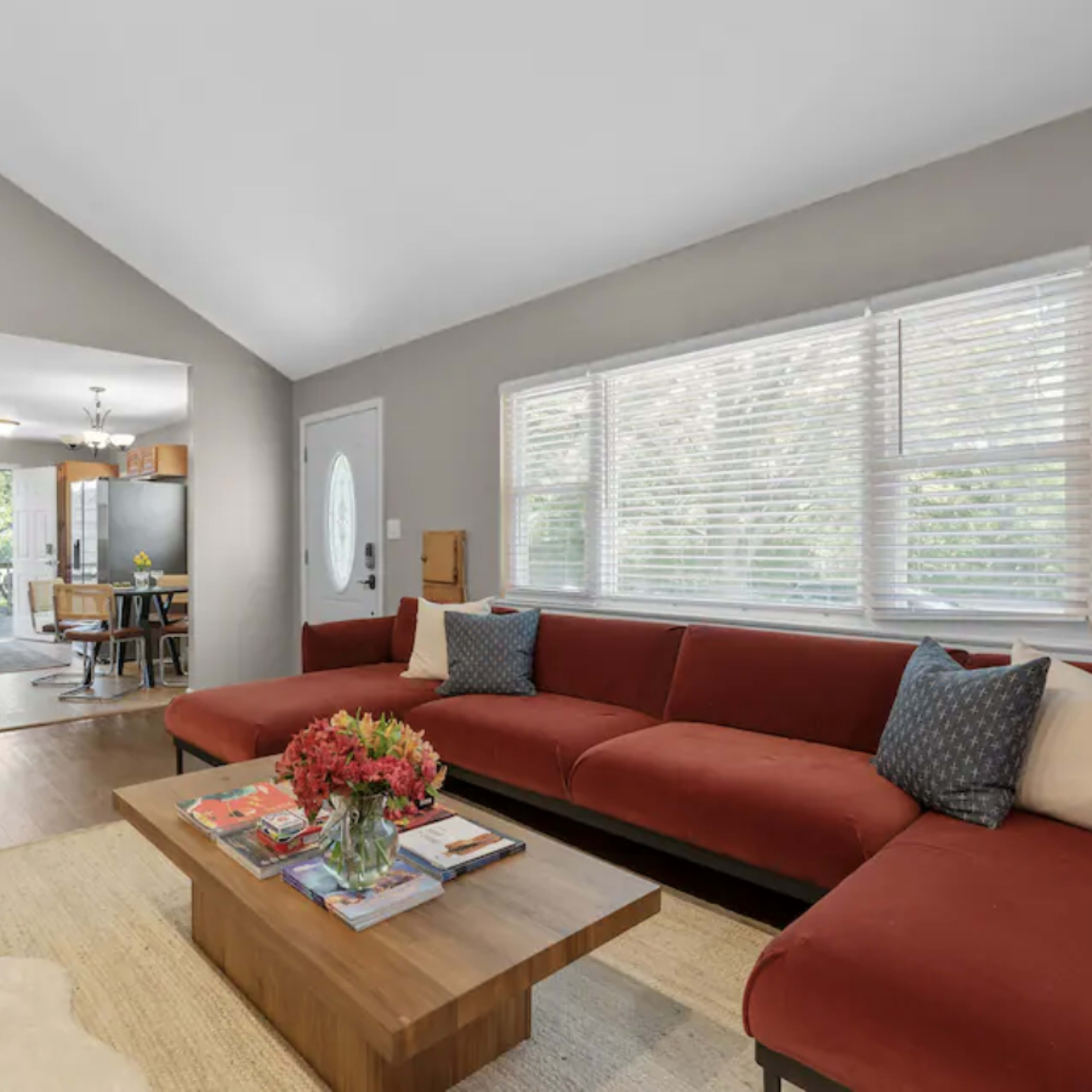 A modern living room features a red sectional sofa, a wooden coffee table with magazines, and large windows allowing natural light into the space.