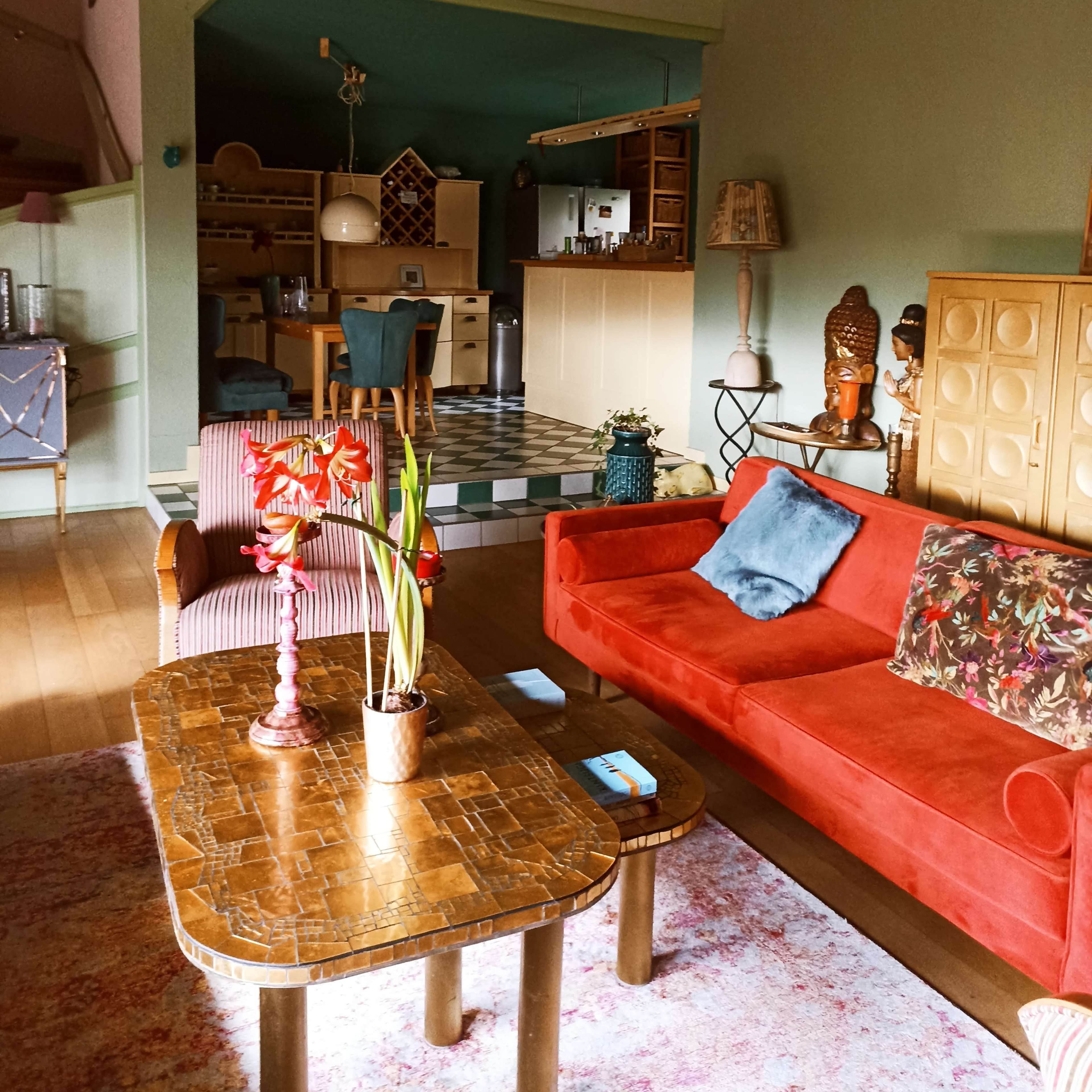 A cozy living room featuring a red couch, a patterned coffee table, and a kitchen area visible in the background with a checkered floor.