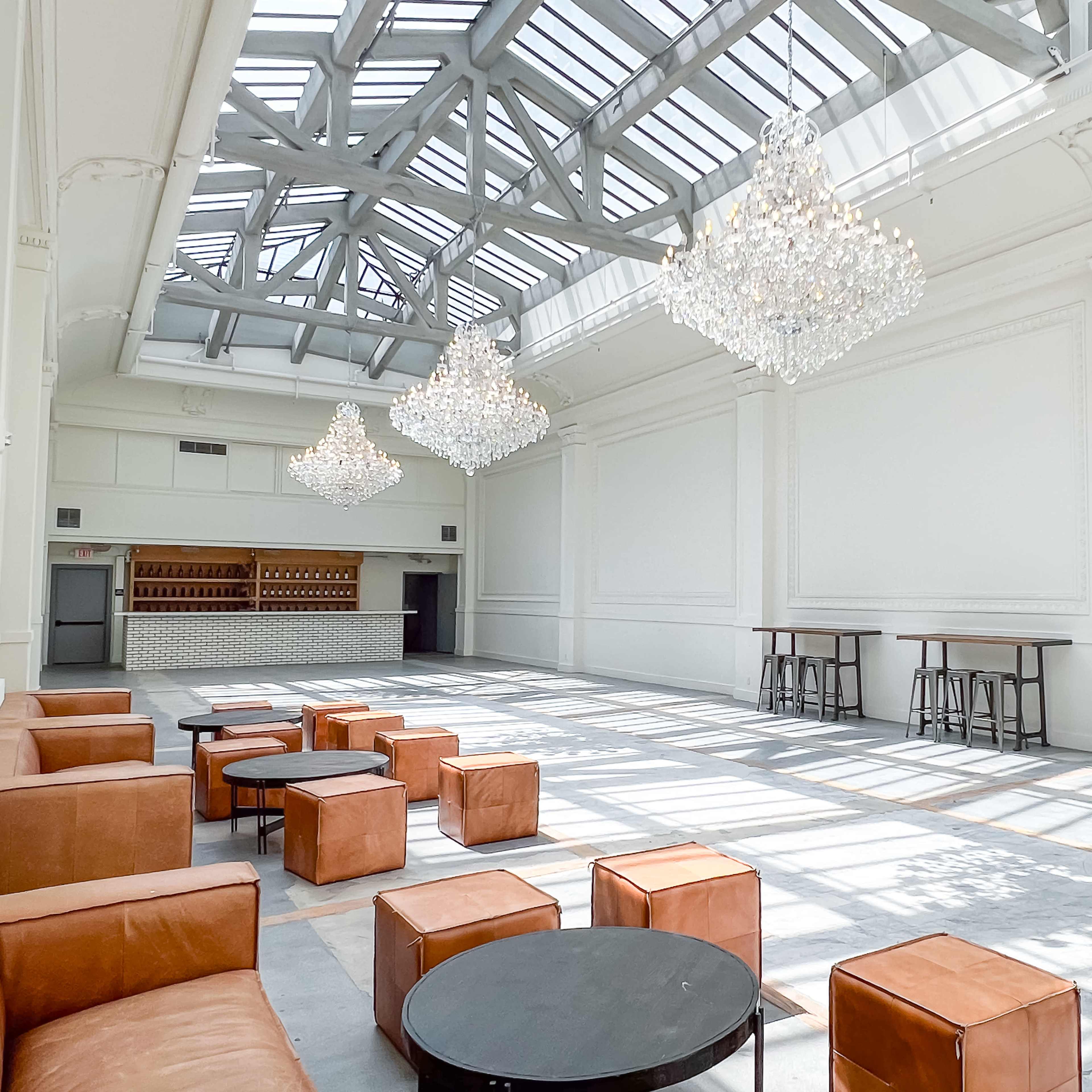 A spacious, well-lit room with large windows, featuring several crystal chandeliers, leather seating, and a bar area in the background.