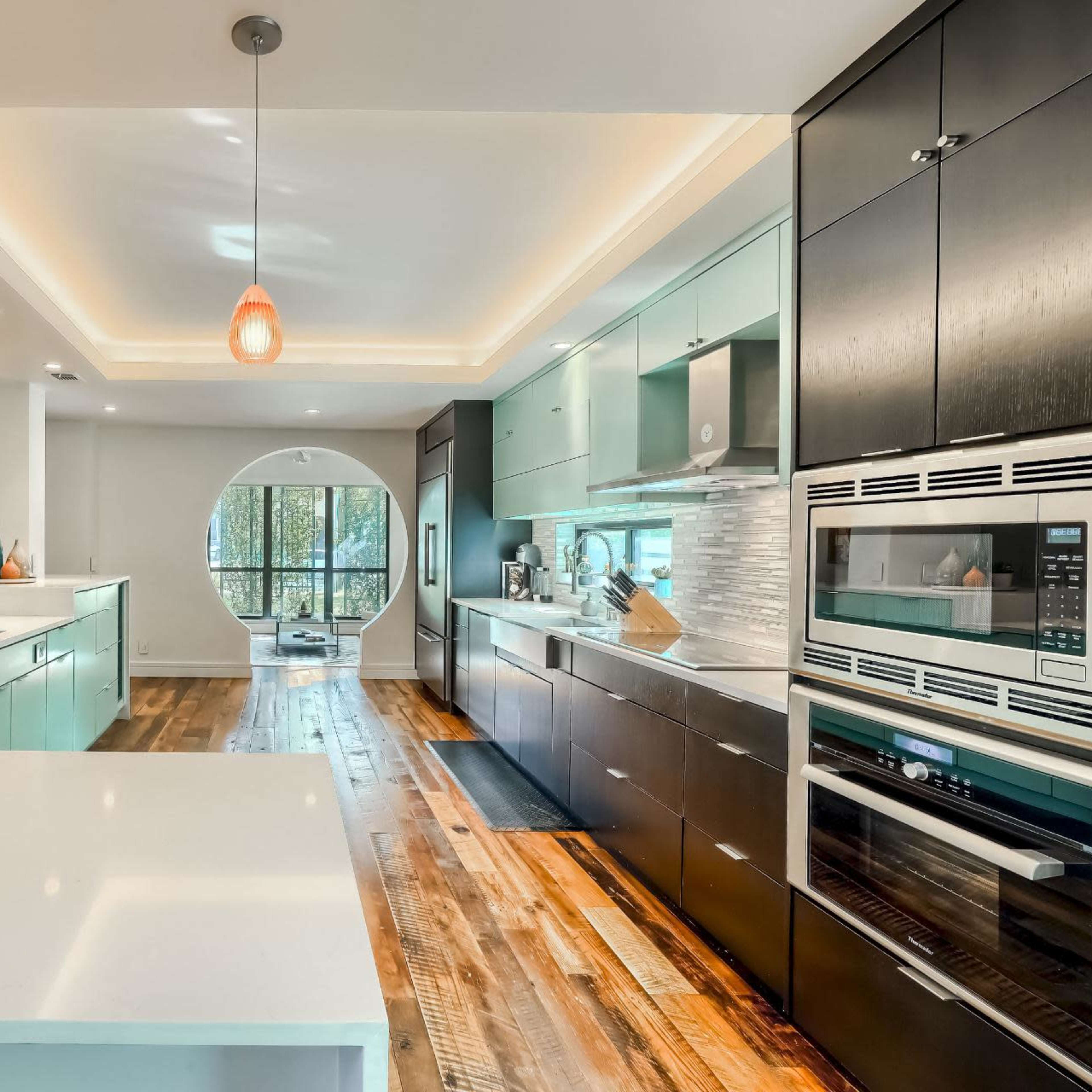 A modern kitchen features a central island with a white countertop, mint green cabinetry, and a circular window that overlooks a wooded view.