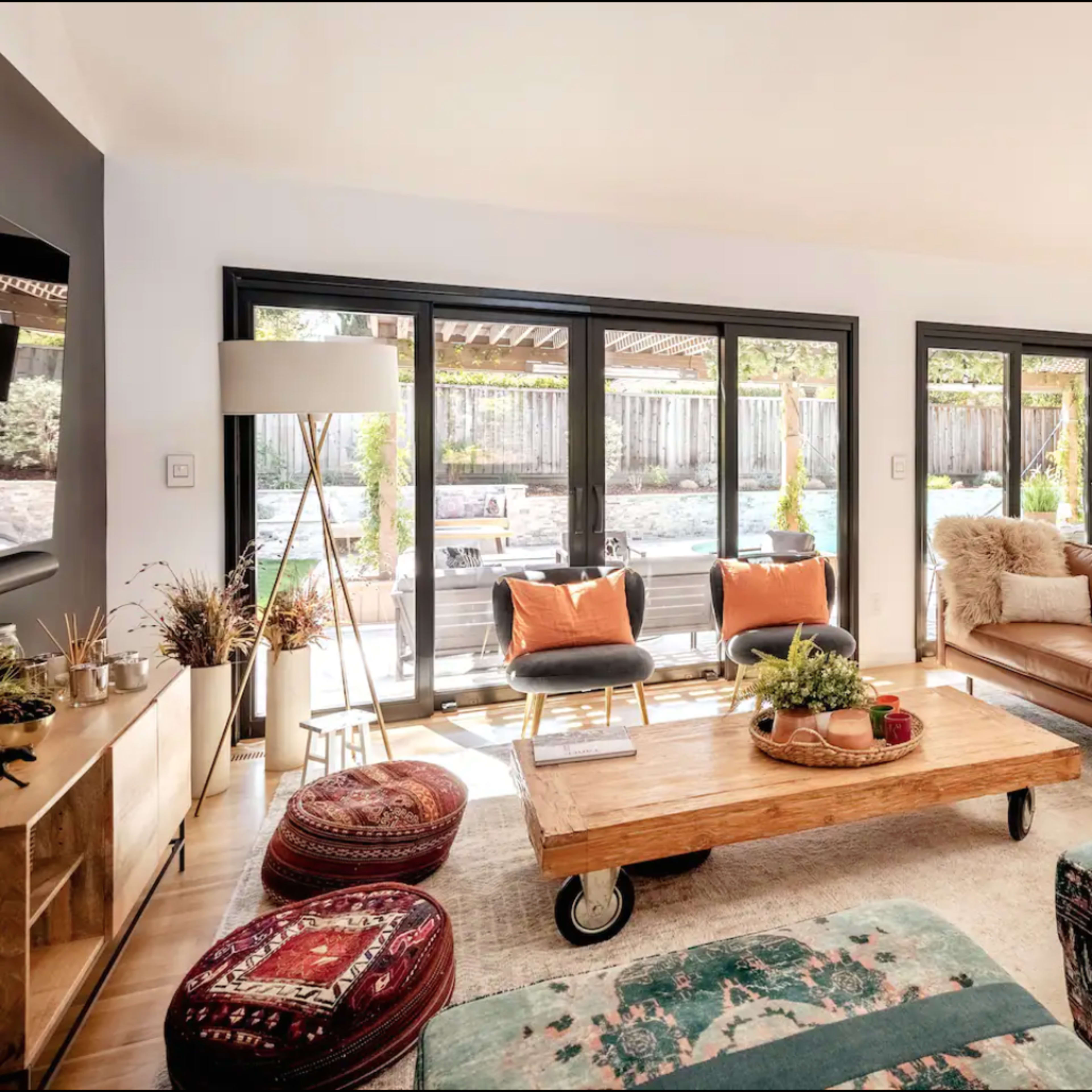 A modern living room features a brown leather sofa, an oversized wooden coffee table on wheels, and large glass doors that open to a backyard with a pool.