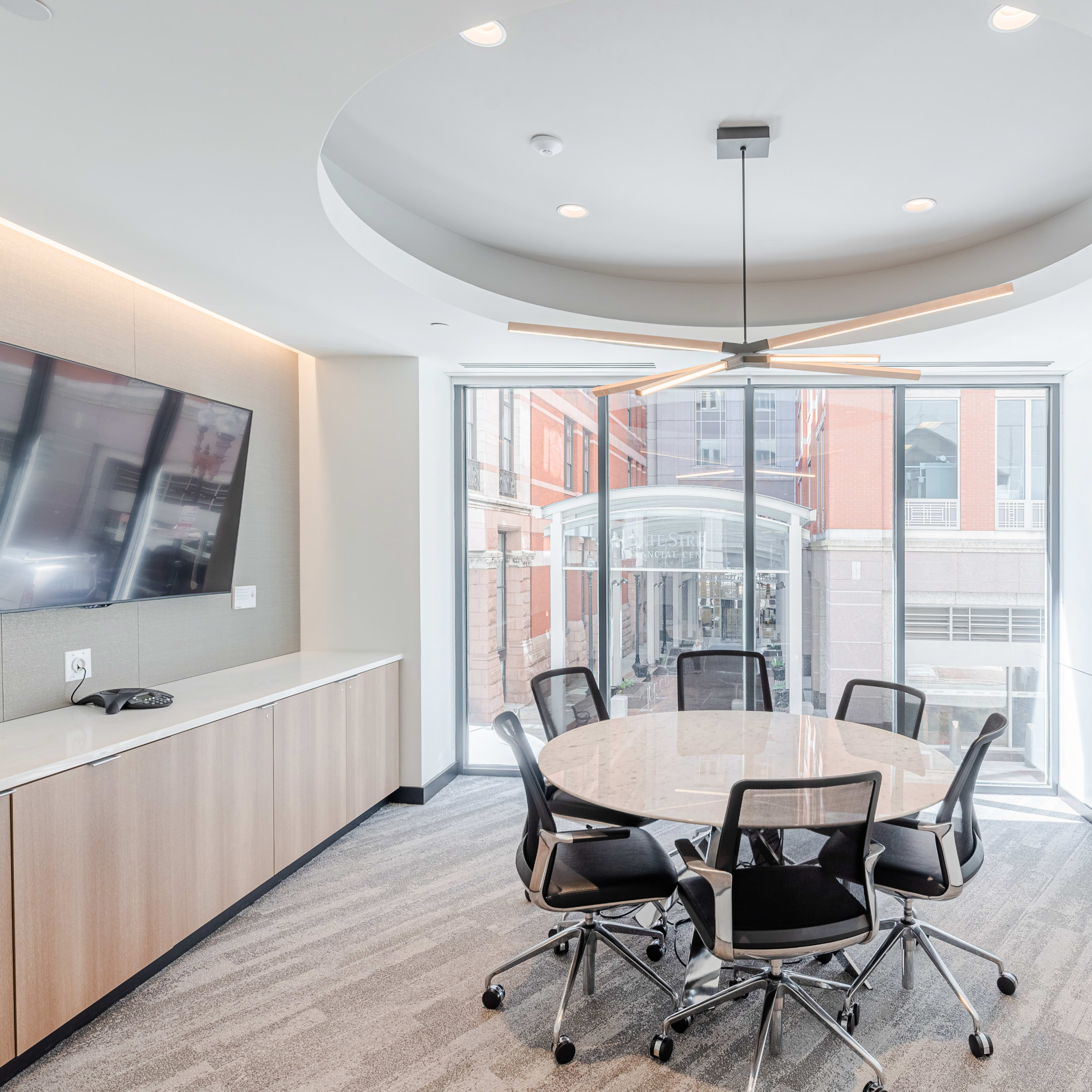 A modern conference room features a round table surrounded by black chairs, a wall-mounted TV, and large windows overlooking an outdoor area.