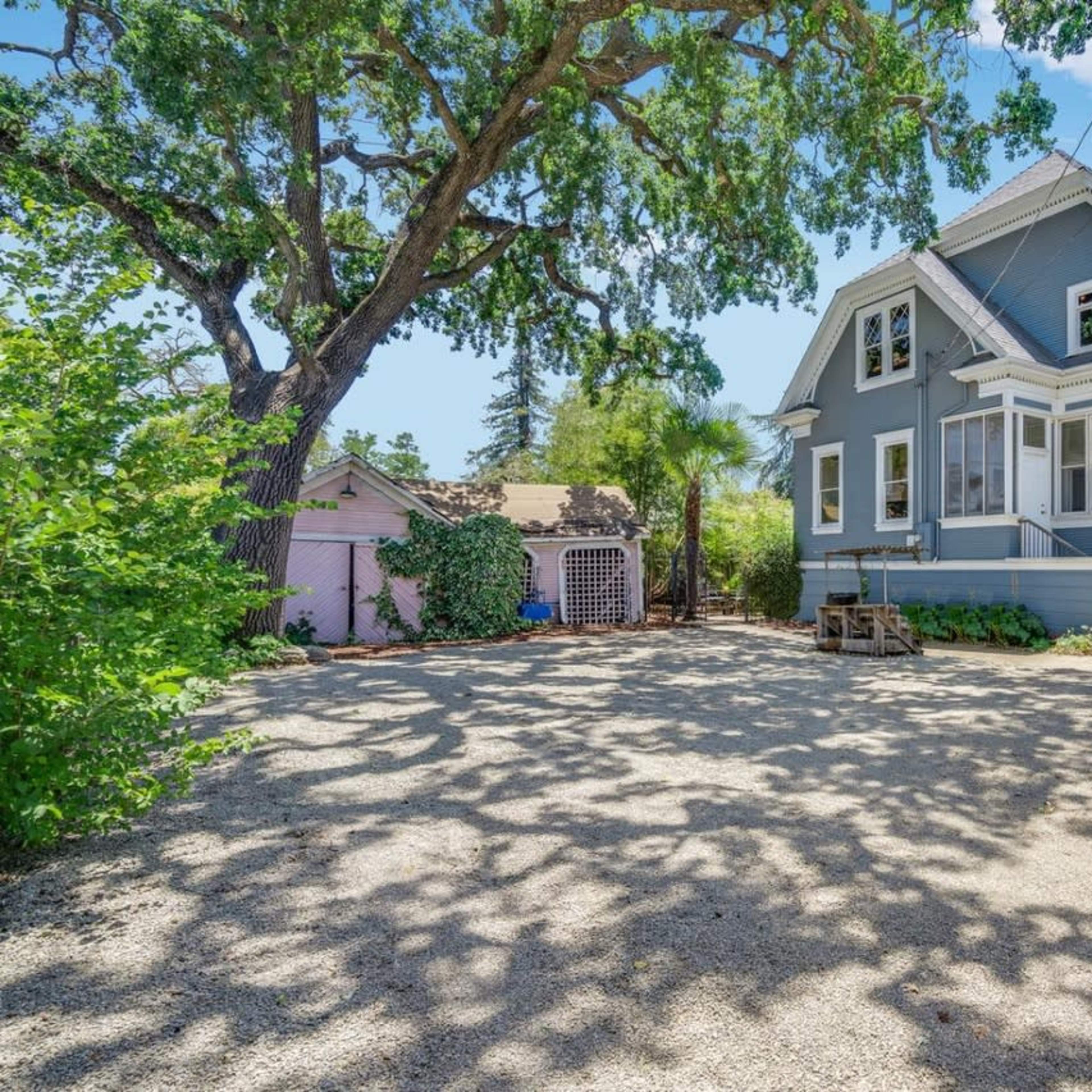 A large, two-story blue house with a covered porch is situated next to a gravel driveway and surrounded by lush greenery and trees.